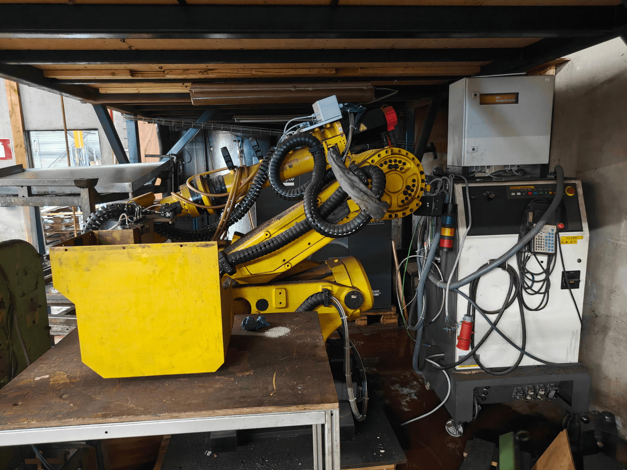 Vue de face d'un bras de robot FANUC S-430i W, de couleur jaune, avec son panneau de commande, ses câbles et un composant jaune posé sur un établi.