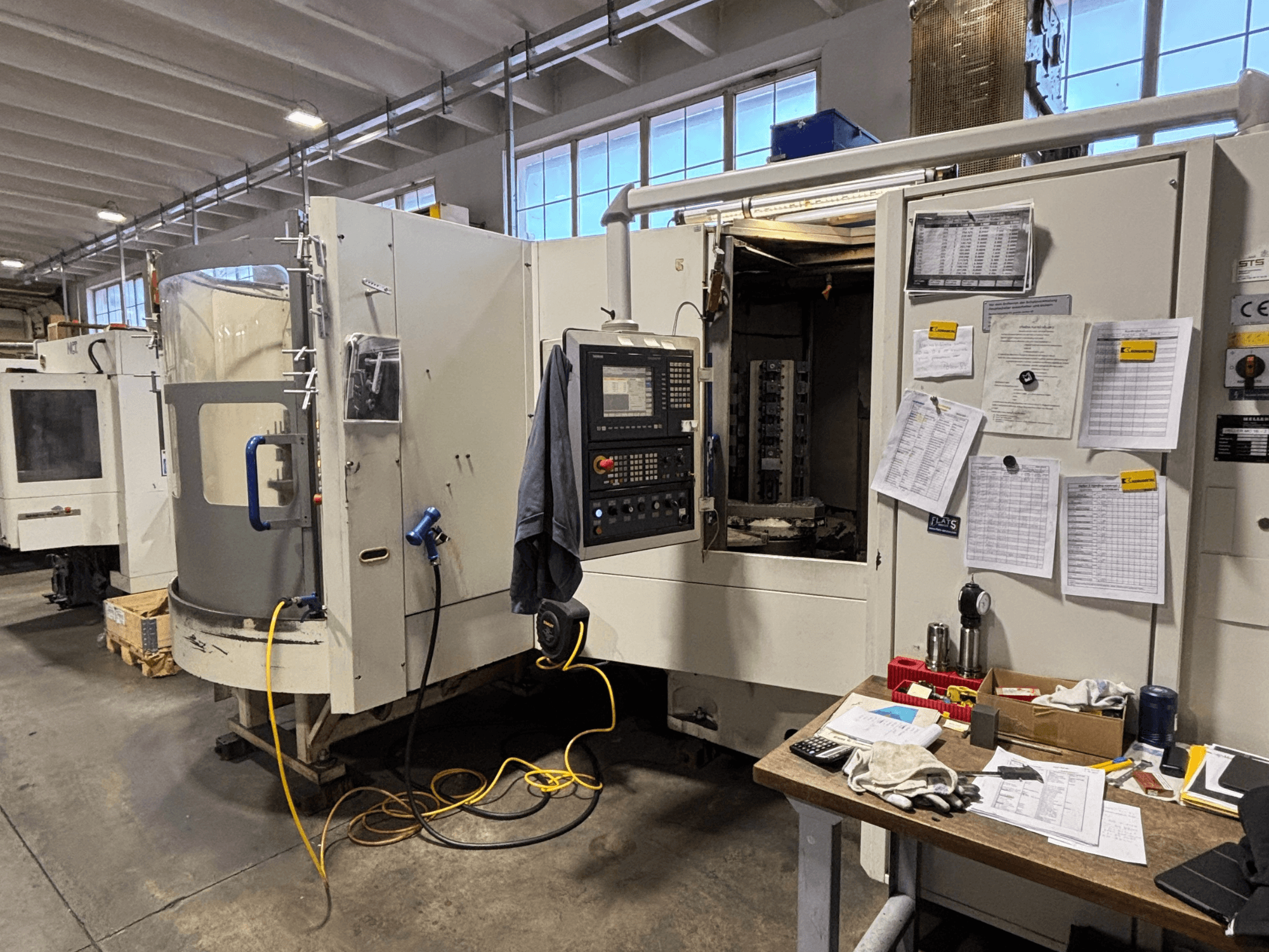 Machine à commande numérique dans un atelier, vue de face, avec panneau de commande et documentation sur le côté ; outils et matériaux sur la table.