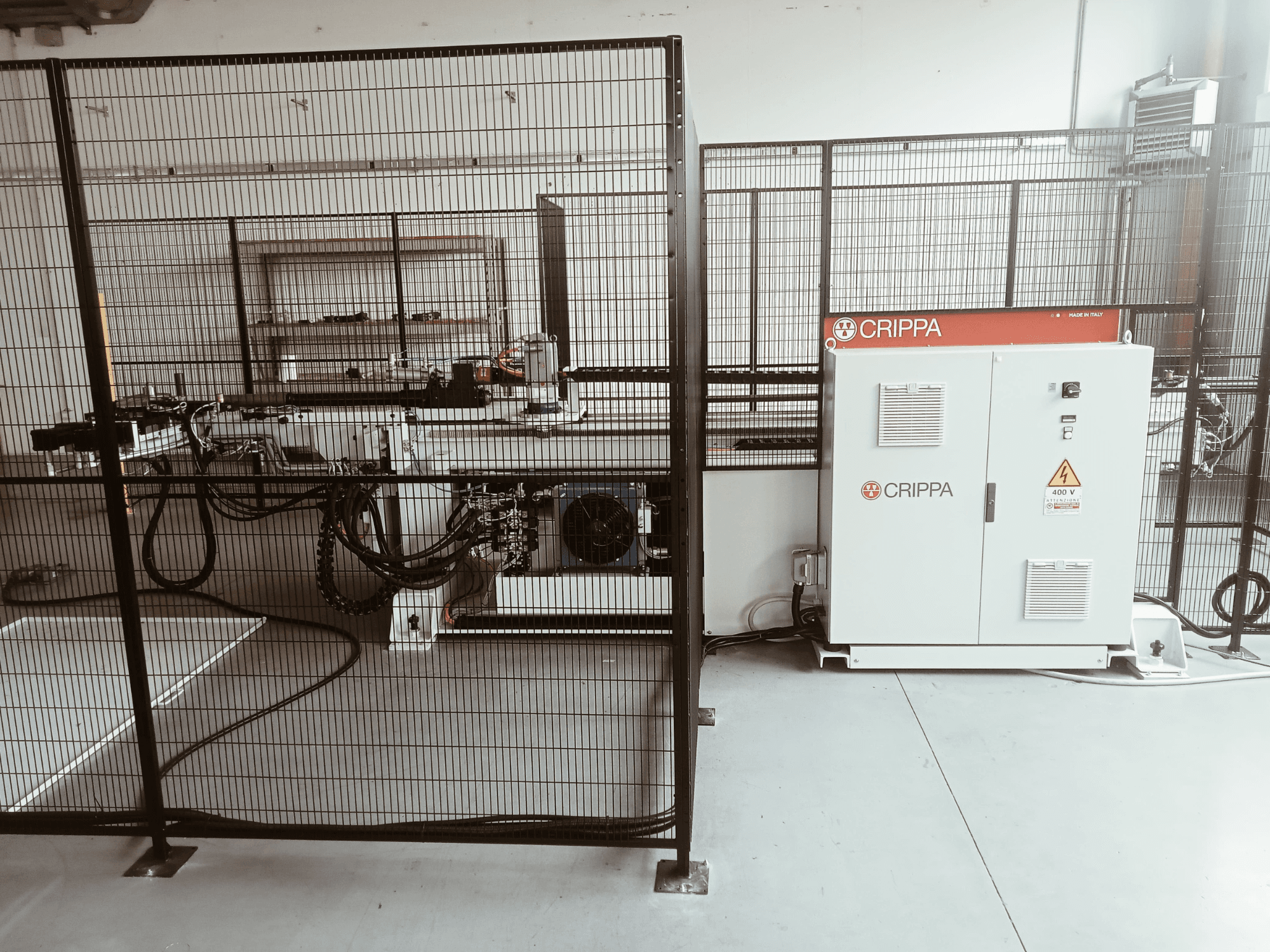 Machine CRIPPA blanche derrière une cage de sécurité, avec des tuyaux et des composants visibles, vue latérale, environnement industriel.