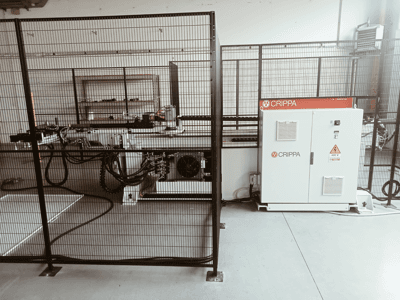 Machine CRIPPA blanche derrière une cage de sécurité, avec des tuyaux et des composants visibles, vue latérale, environnement industriel.