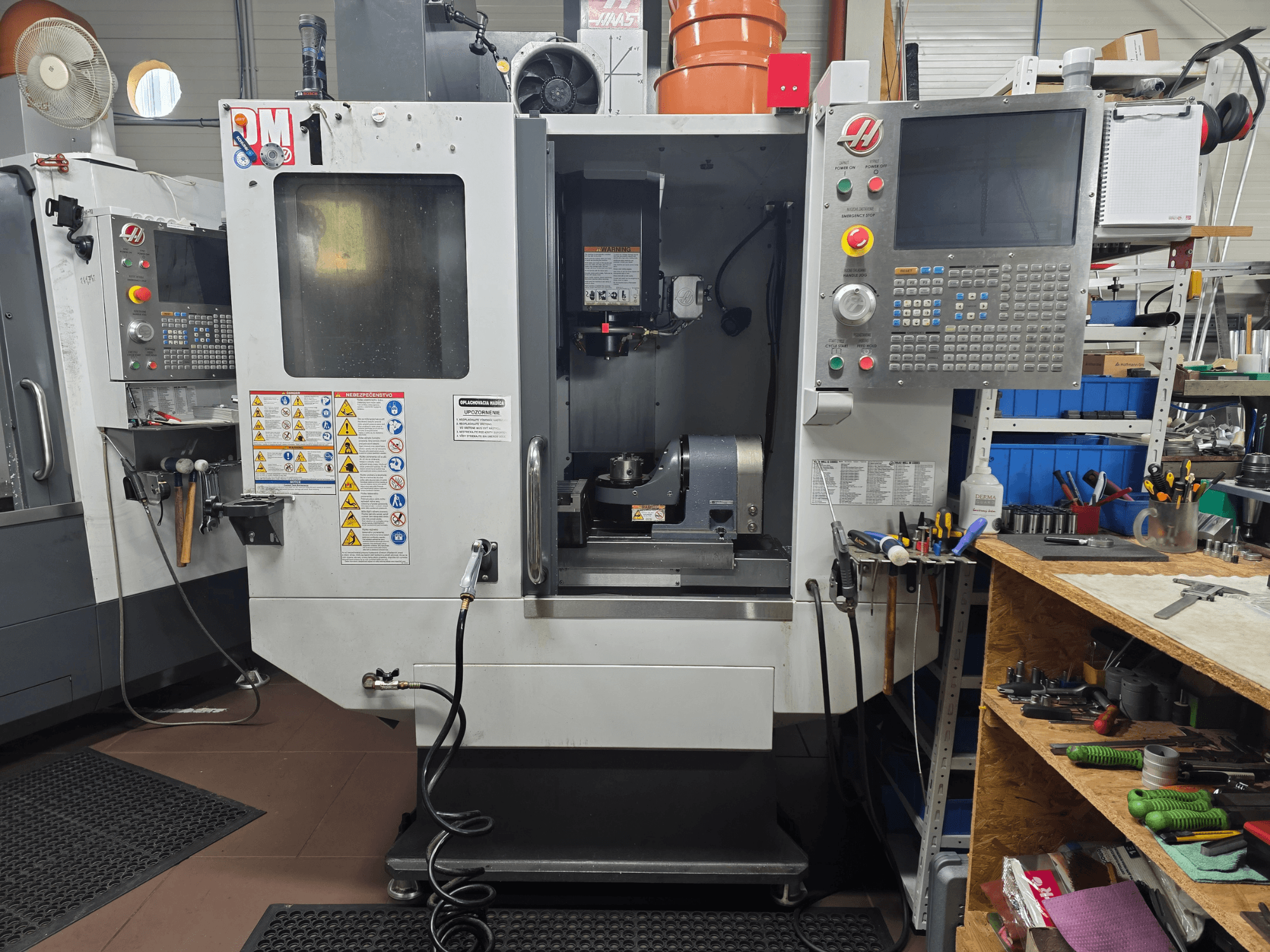 Machine CNC Haas modèle VF-2, vue de face, montrant les commandes, la zone de la broche, les étiquettes de sécurité et les outils sur un établi proche.