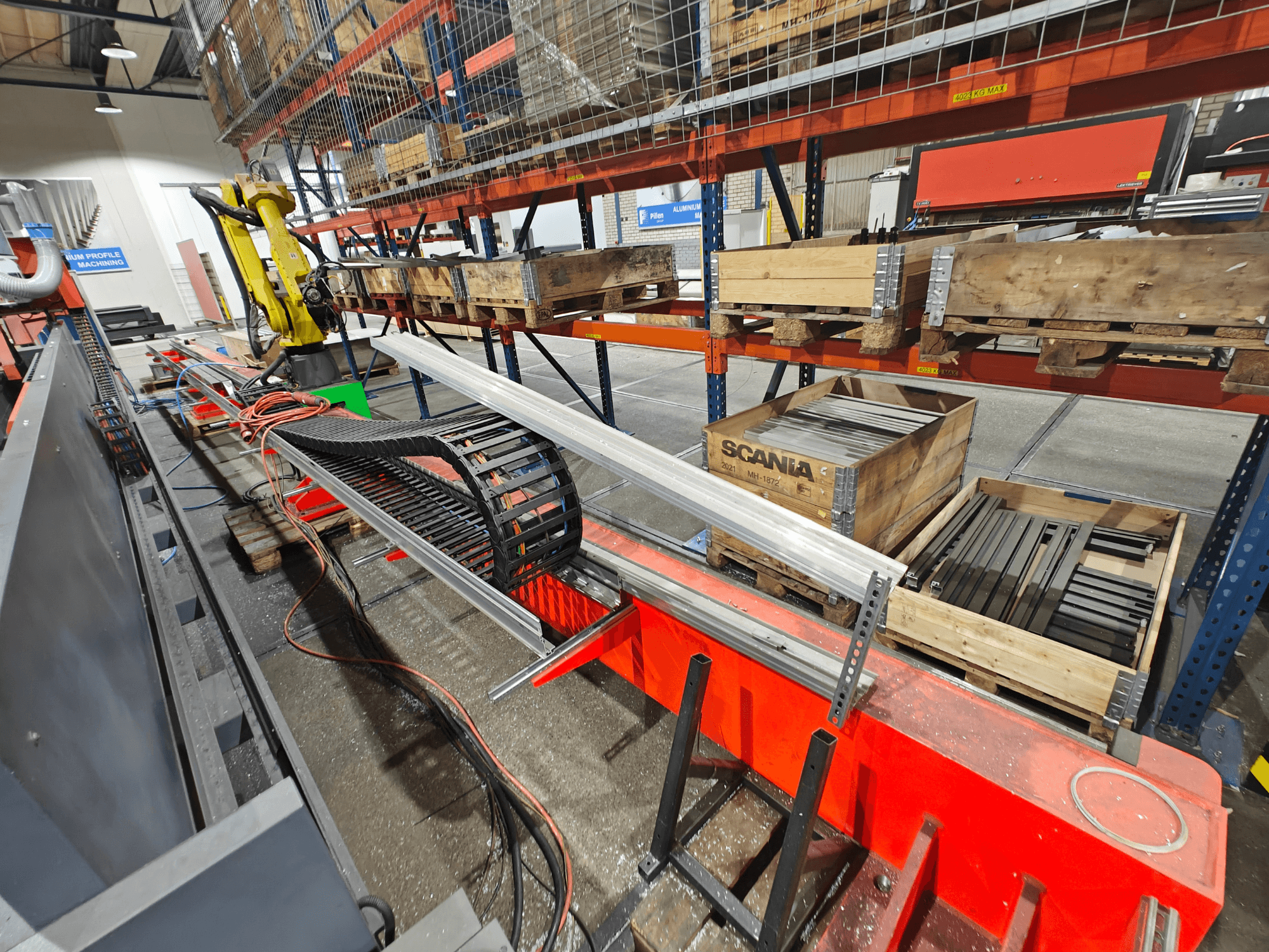 Bras robotisé sur une station de travail, câbles orange et noirs visibles, avec des caisses Scania et des profilés en aluminium dans une usine.