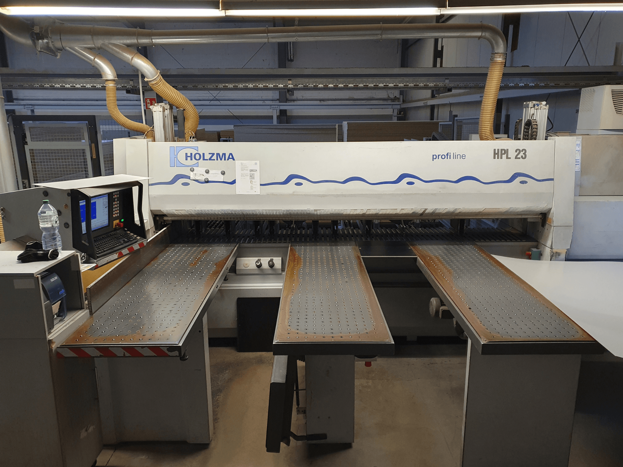 Machine à découper HOLZMA HPL 23 vue de face, avec trois tables perforées, un terminal d'ordinateur et une bouteille d'eau à proximité.