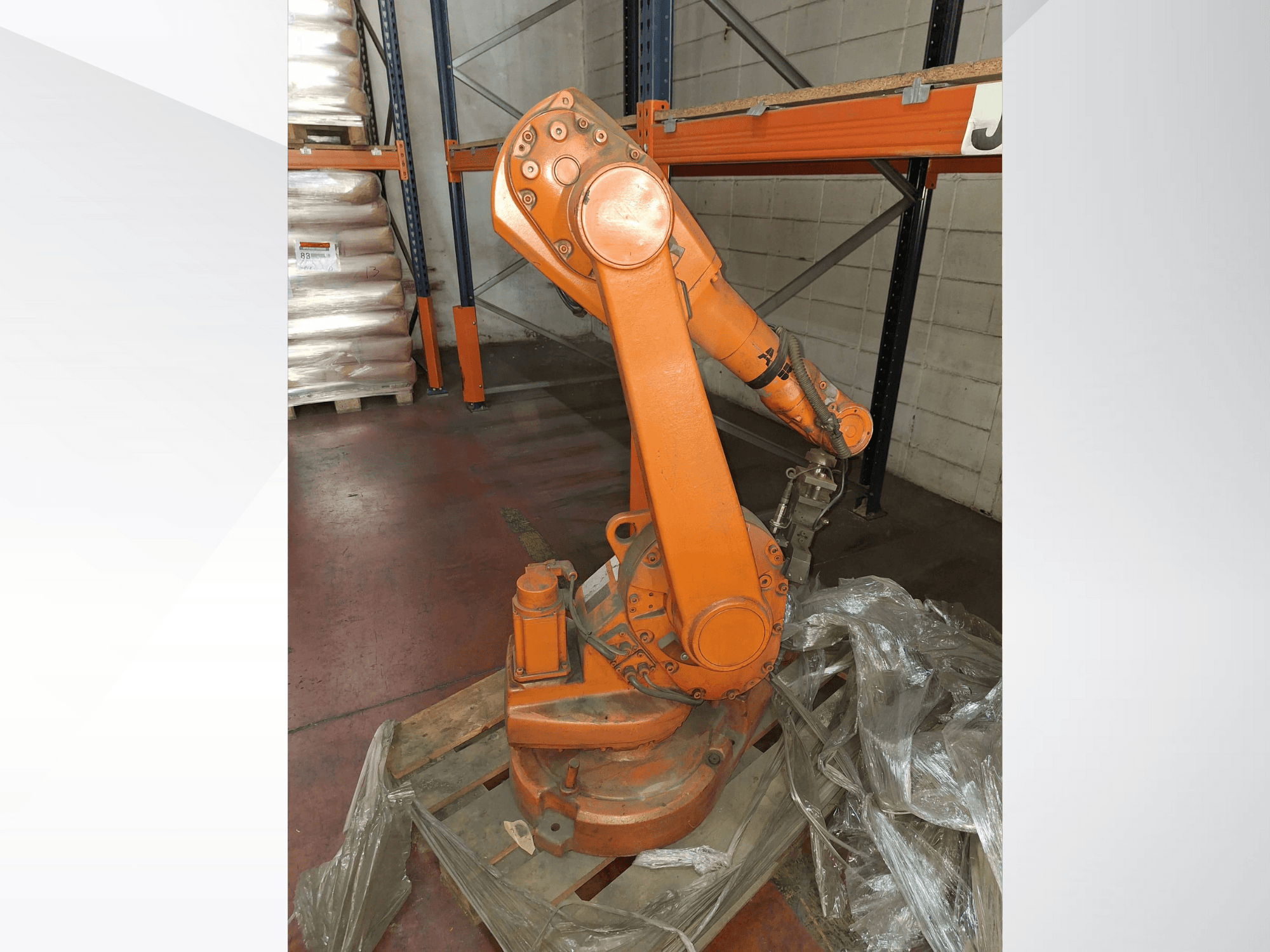 Bras robotique industriel de couleur orange, vue de face sur une palette en bois, avec câble attaché et film plastique de protection autour de la base.