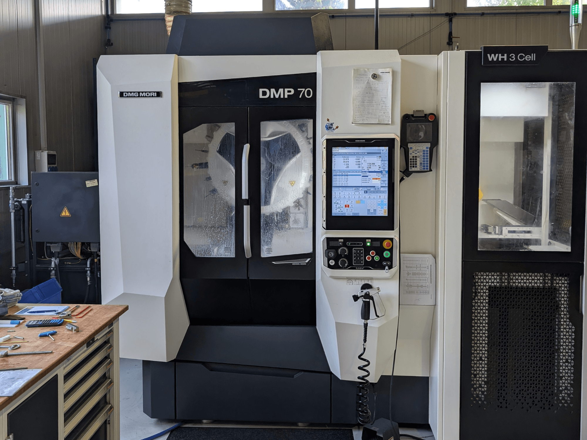 Centre d'usinage vertical DMG MORI DMP 70, vue de face avec le panneau de commande et l'écran de contrôle, entouré d'outils d'atelier.