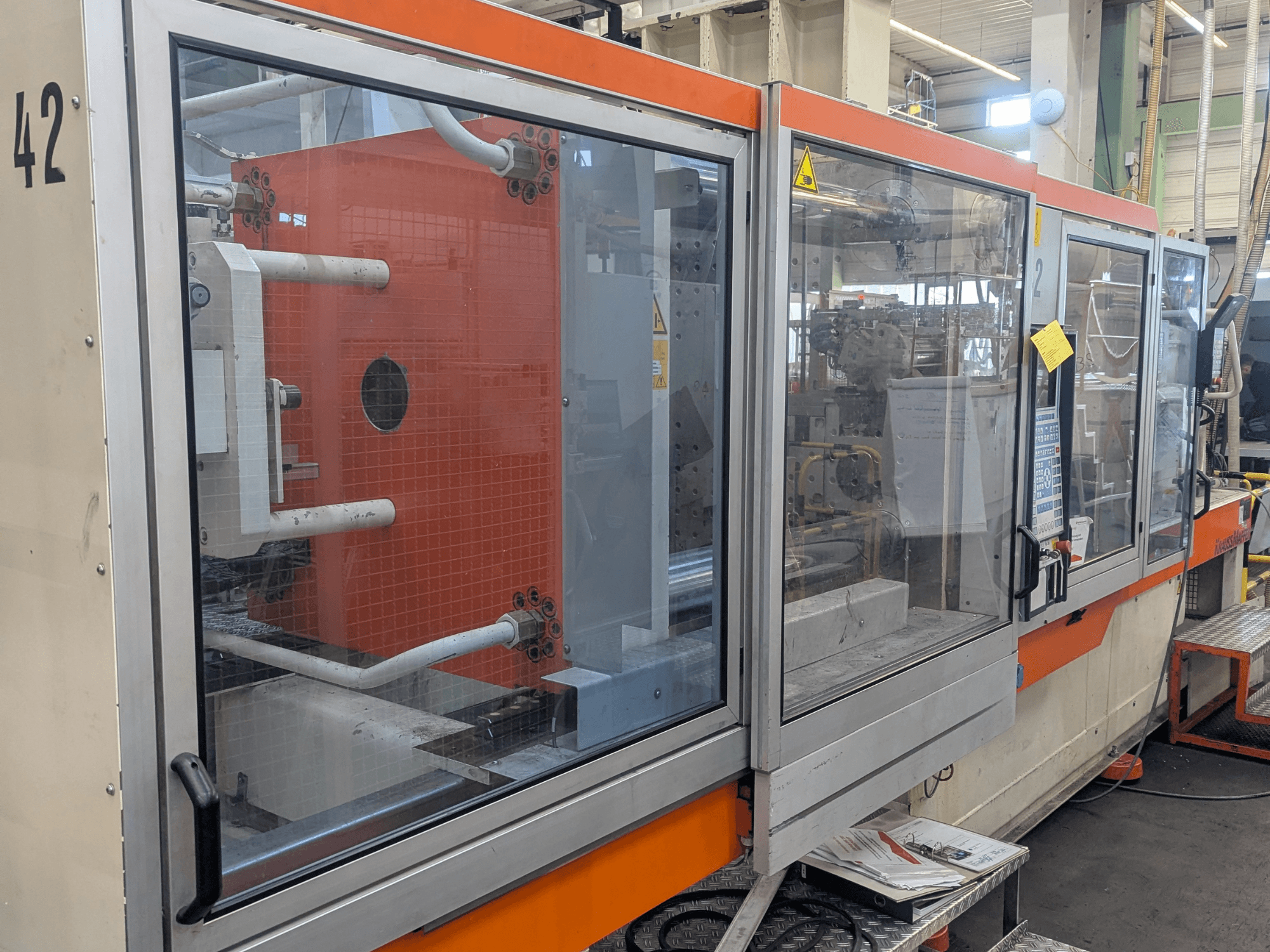 Machine de moulage par injection hydraulique Krauss Maffei KM 350-3000, vue latérale montrant les commandes et les composants internes derrière un panneau en verre.
