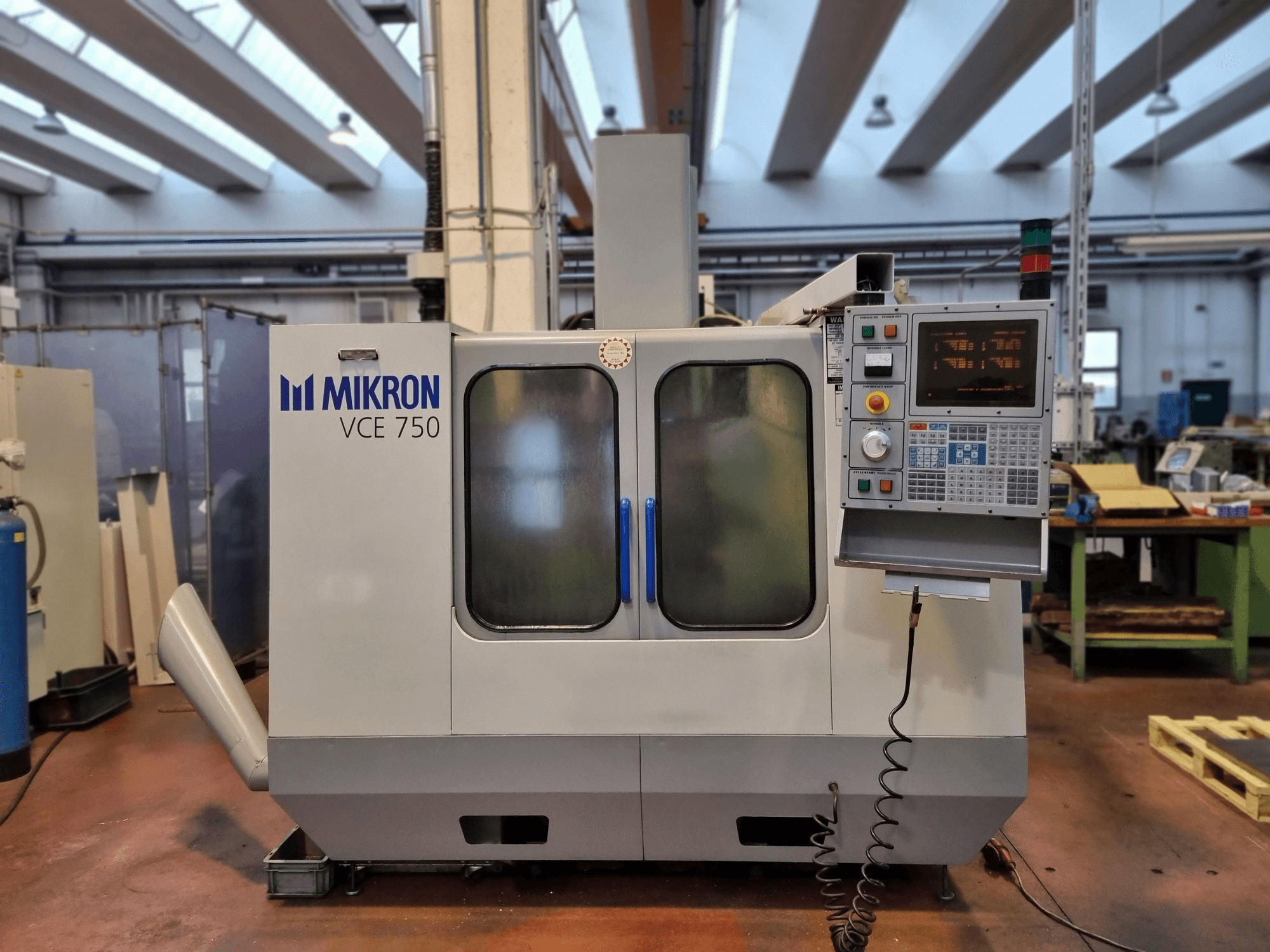Machine CNC Mikron VCE 750 blanche dans un atelier, vue de côté, avec un panneau de commande numérique et de grandes portes vitrées.