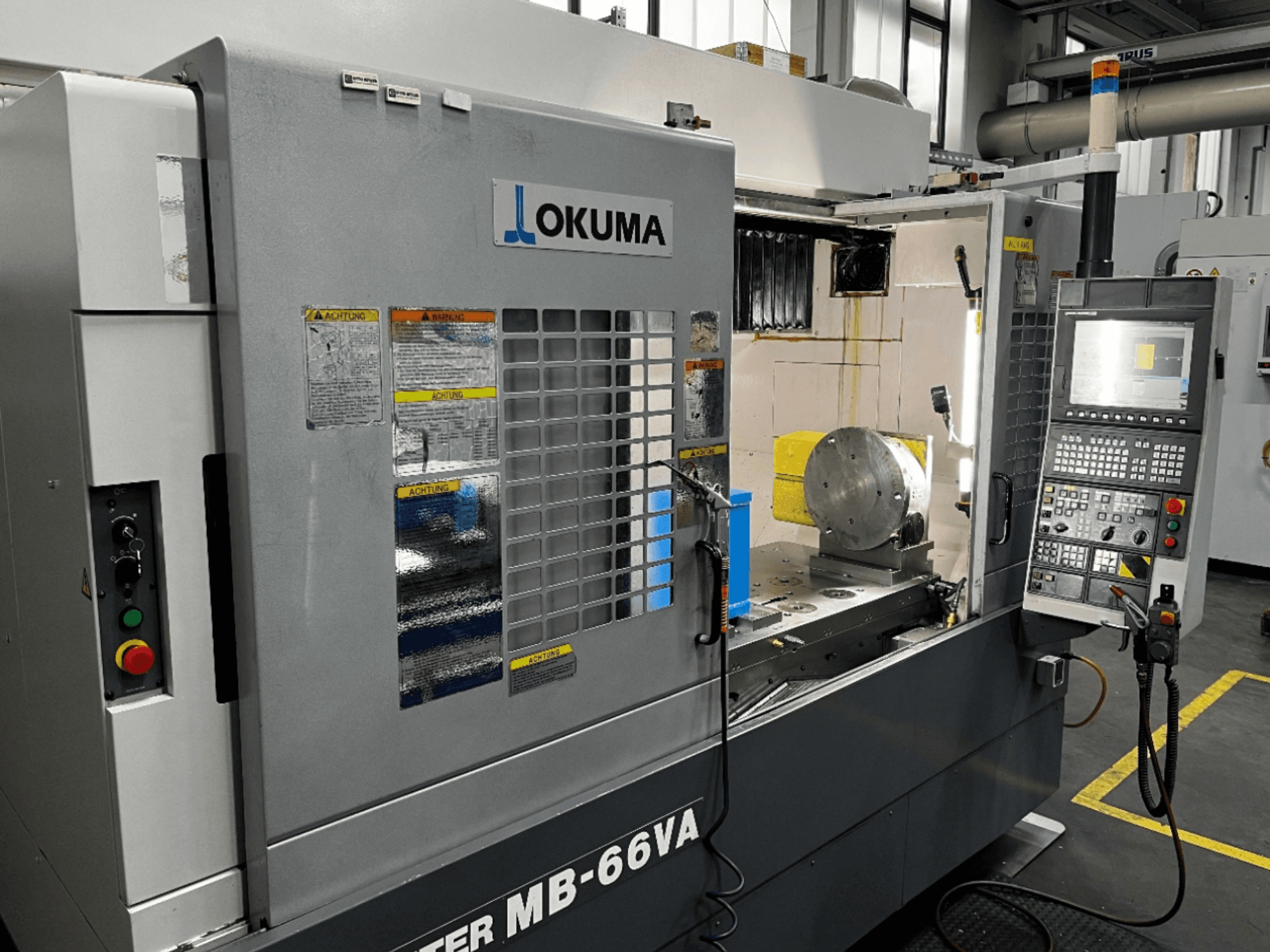 Vue avant du centre d'usinage vertical Okuma MB66-VA montrant le panneau de commande, la pièce à usiner et l'intérieur de la machine avec les étiquettes de sécurité.