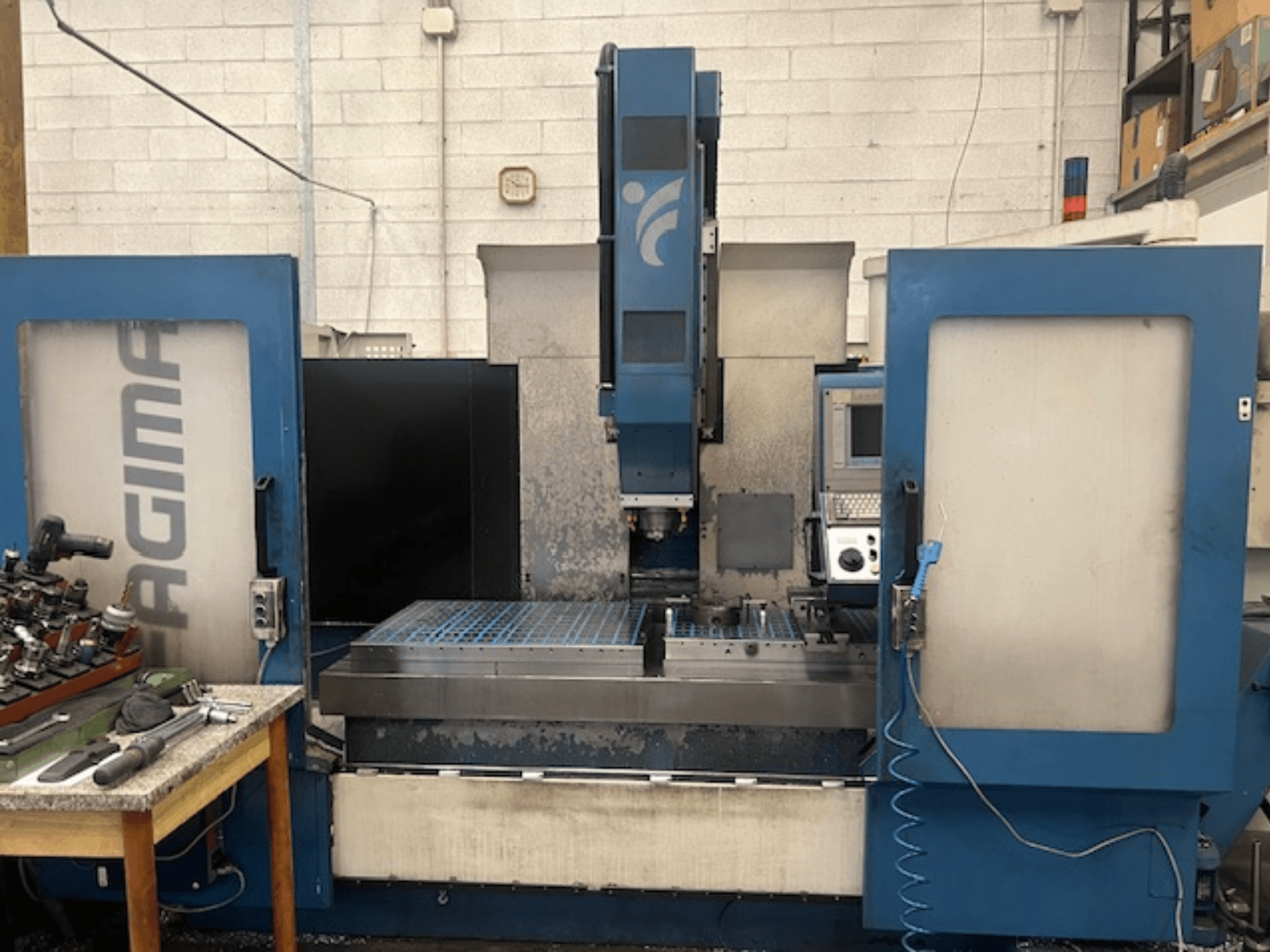 Vue de face d'une machine CNC JAGIMA avec un grand espace de travail en métal, un panneau de commande visible et des outils posés sur une table voisine.