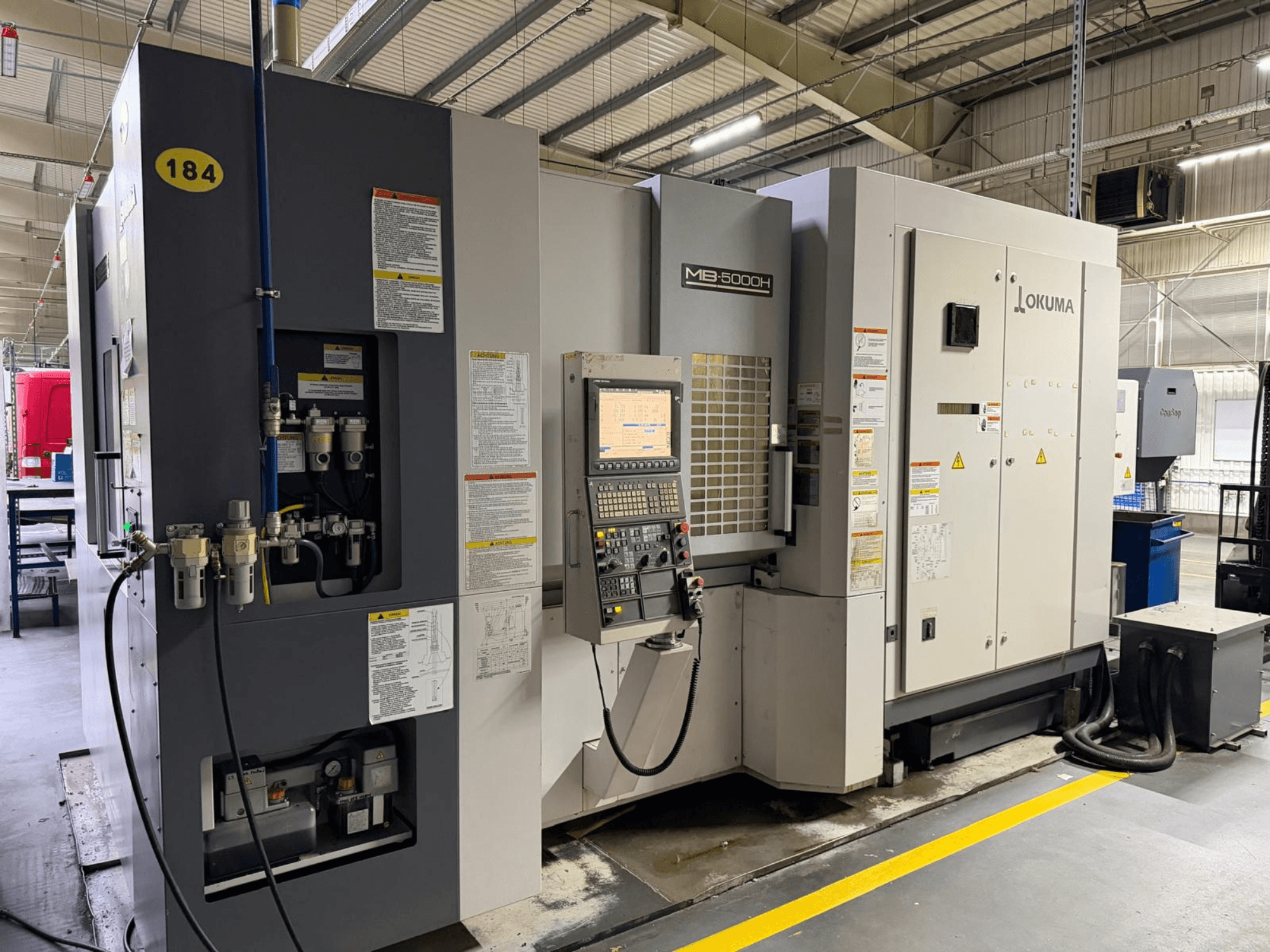 Vue avant du centre d'usinage horizontal Okuma MB 5000H, avec les commandes, l'étiquetage et les connexions hydrauliques.