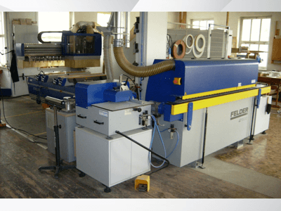 Plaqueuse de chants FELDER G500 dans un atelier, vue de profil montrant la structure bleue et jaune avec les commandes et les conduits.