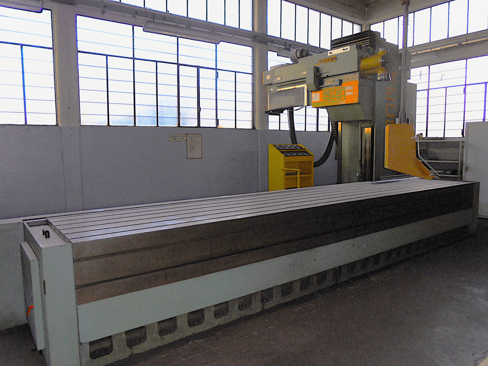 Fraiseuse verticale de marque inconnue dans une usine, vue de côté, avec une grande table à surface rainurée et un panneau de commande.