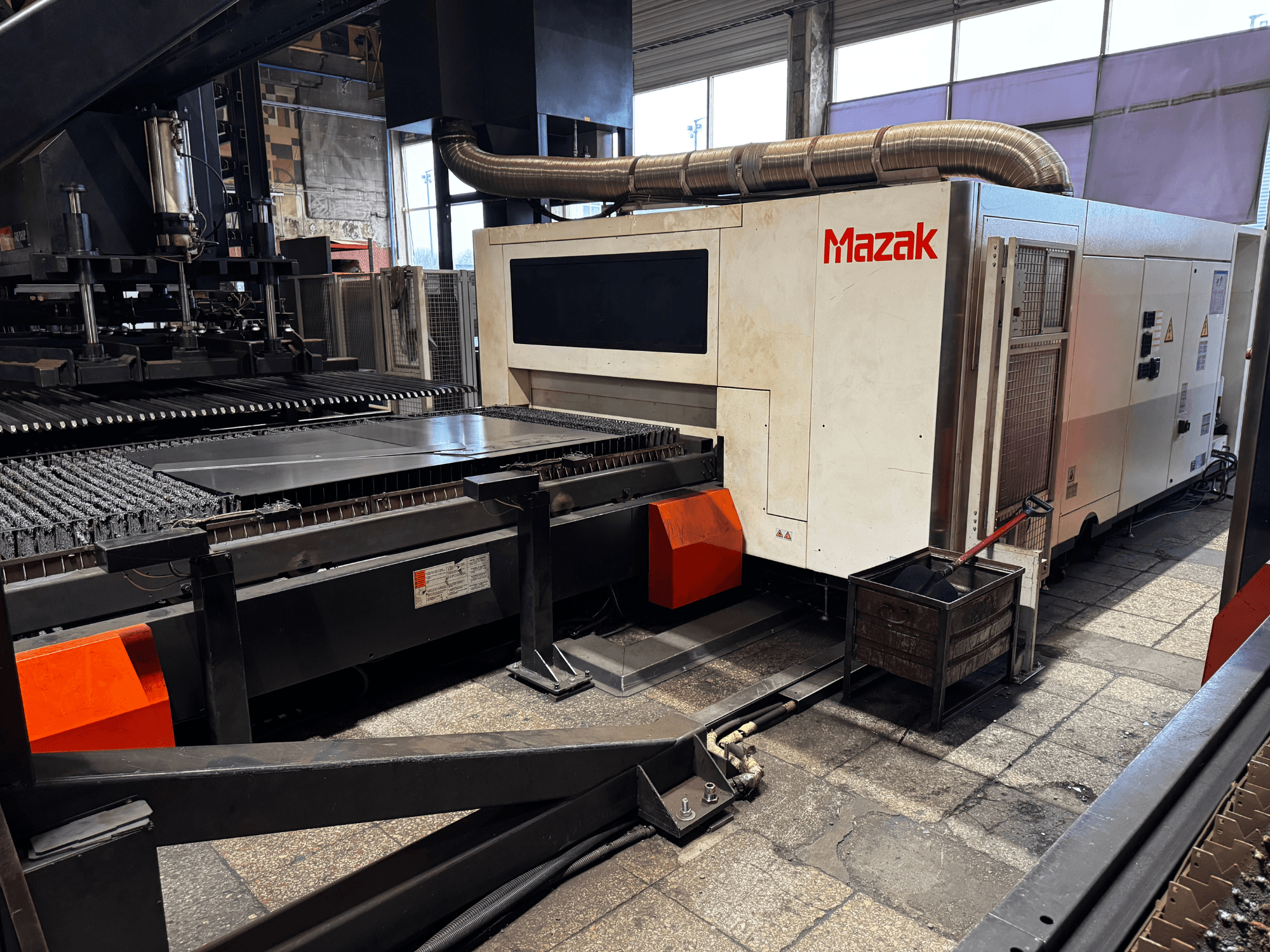Machine de découpe laser Mazak dans un atelier, vue latérale montrant le panneau de commande, le convoyeur et un bac de récupération.