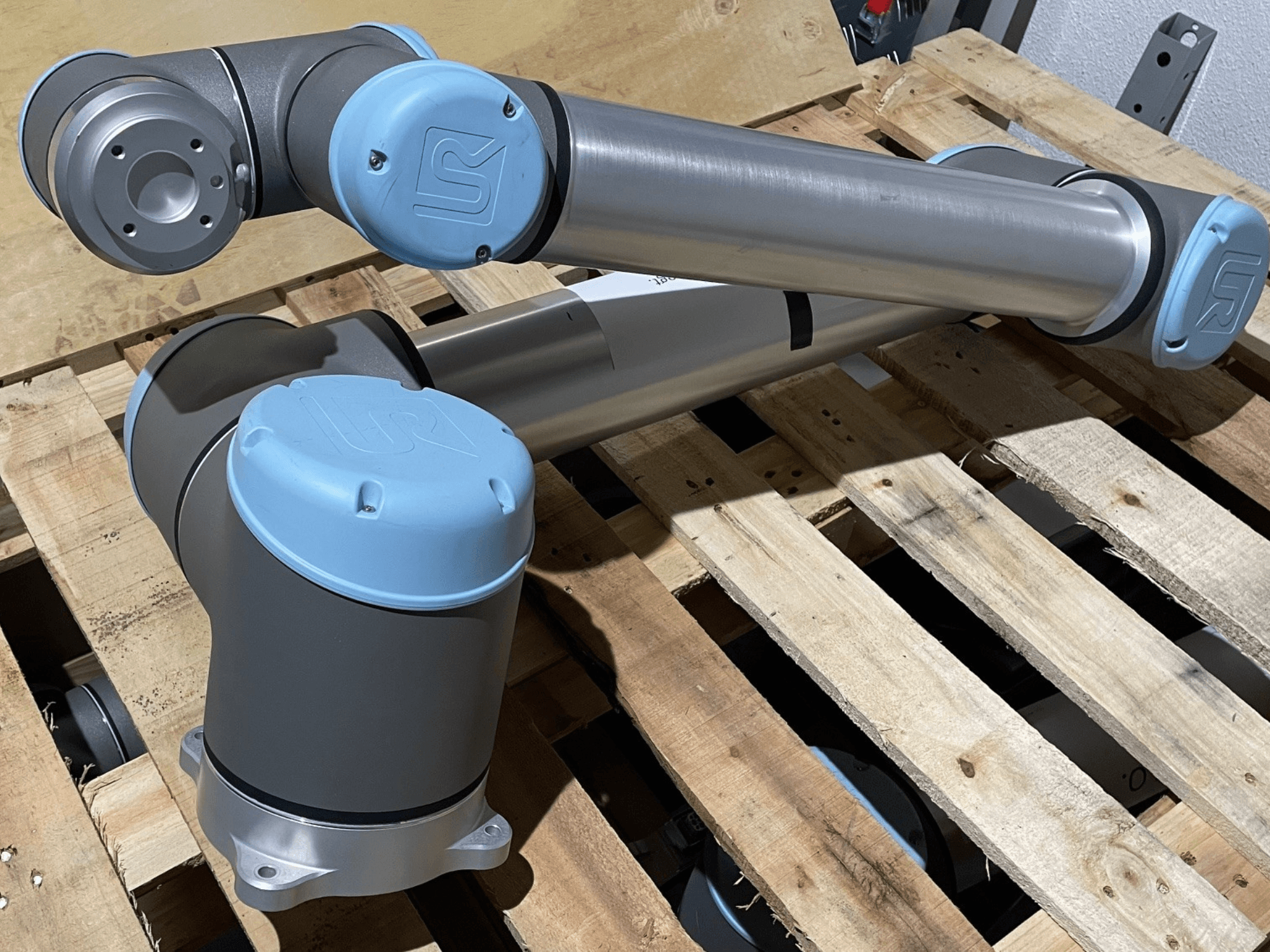 Bras robotique argenté avec embouts bleus, vue de face, montrant les articulations et les détails mécaniques, reposant sur une palette en bois.