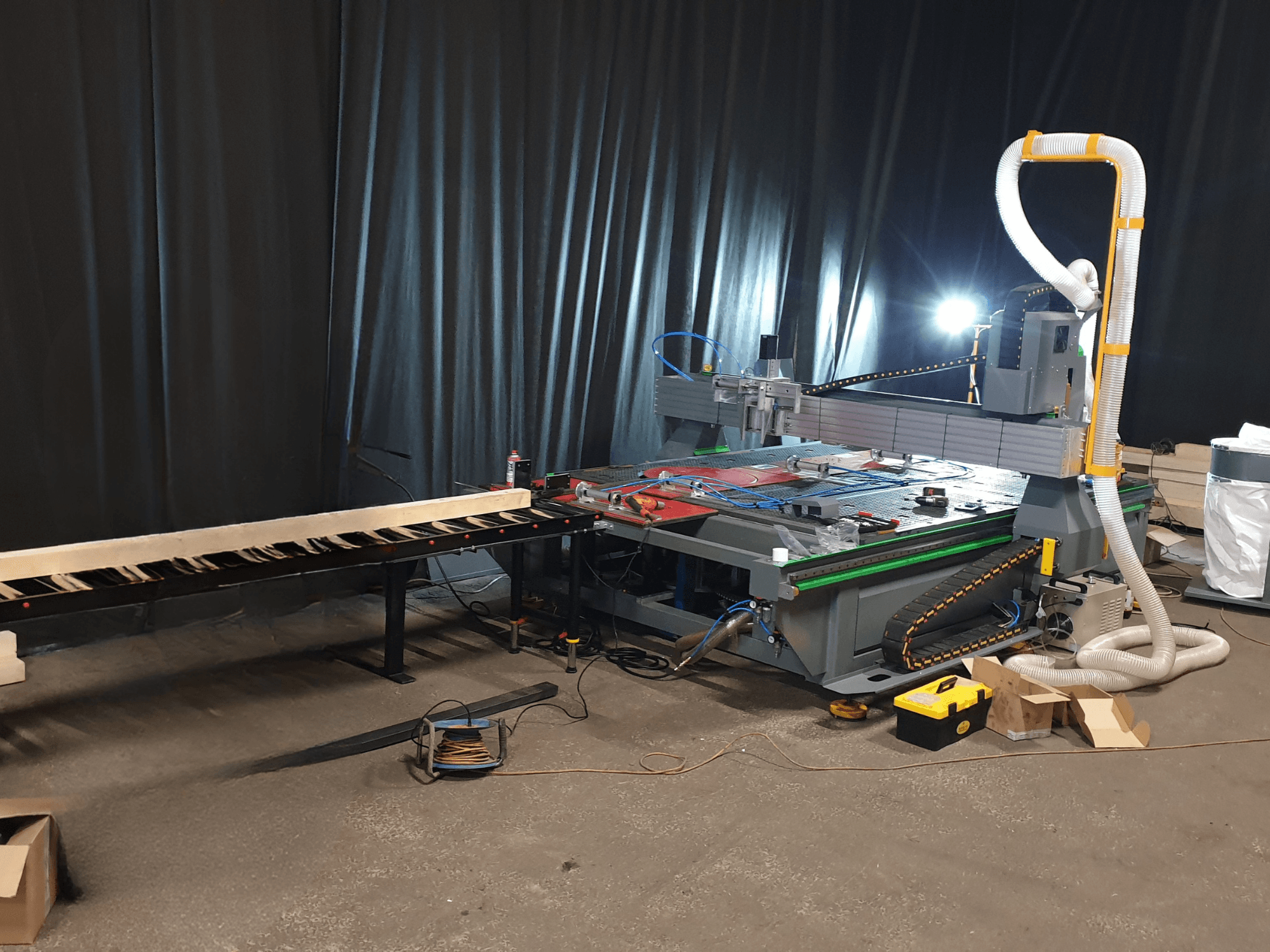 Machine CNC avec convoyeur latéral, outils sur la table et conduit pour le dépoussiérage. Fond noir, vue de face.