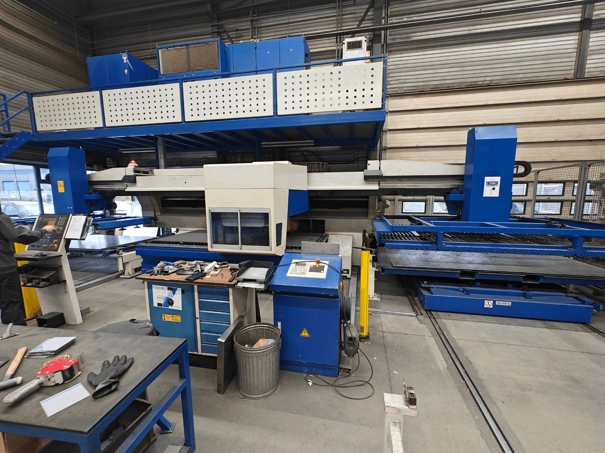 Machine industrielle à commande numérique vue de face, avec une structure bleue et blanche, un panneau de commande et divers outils sur la table de travail.