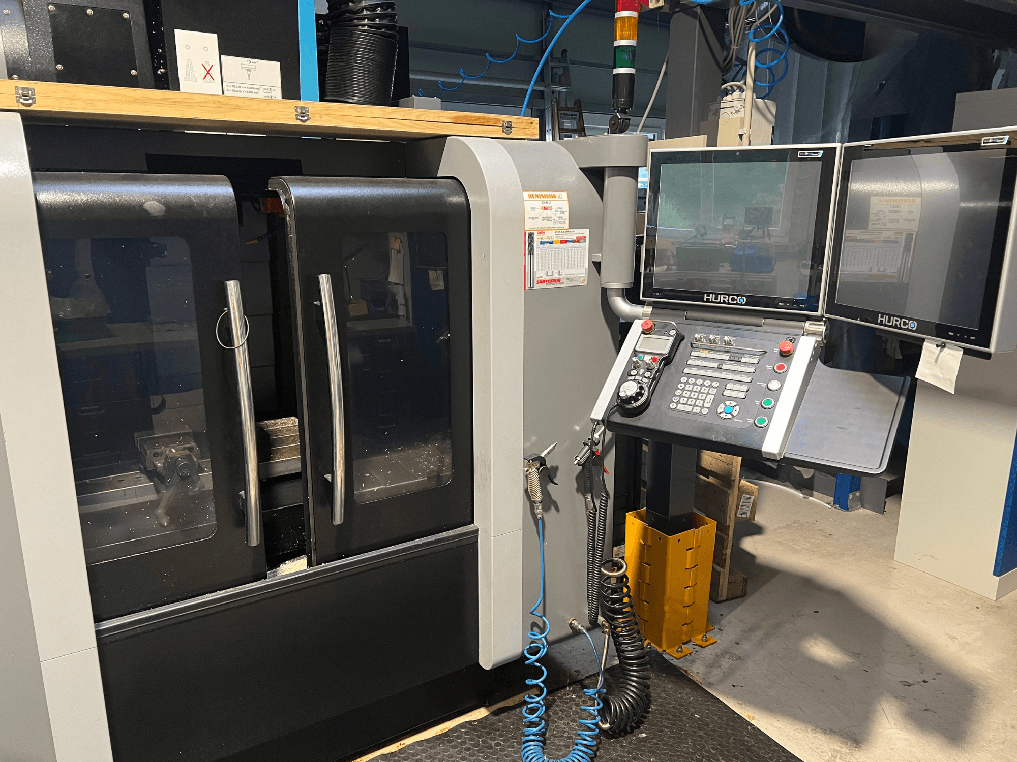 Vue avant de la machine CNC Hurco avec deux moniteurs et un panneau de commande. Les portes sont transparentes et la zone d'usinage est visible à l'intérieur.