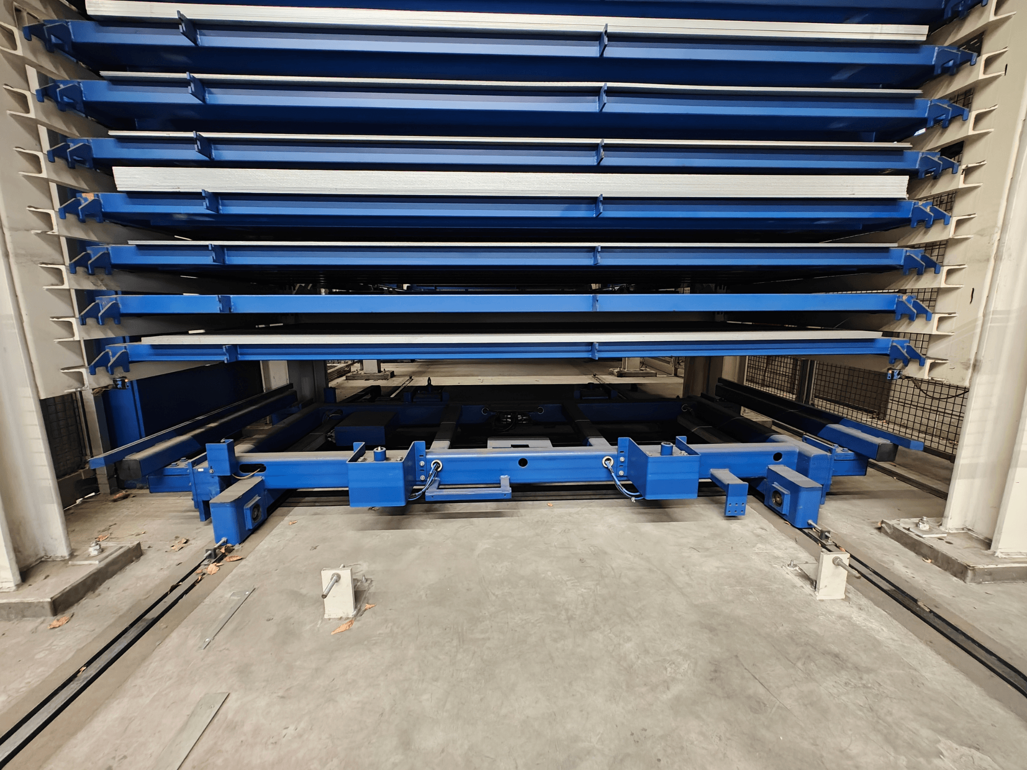 Rayonnage de stockage industriel bleu avec plusieurs étagères horizontales, vu de face. Comprend un mécanisme de levage et des poutres de soutien.