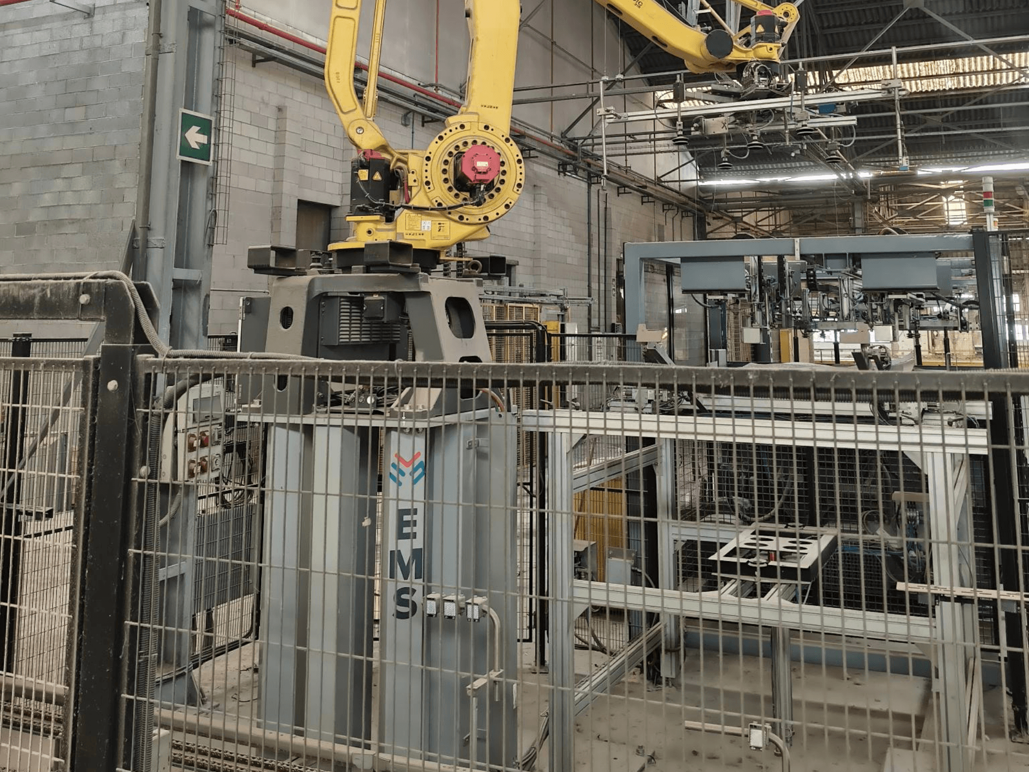 Vue de face du bras robotisé FANUC M-410iC/315 placé au-dessus d'un outil EMS, avec, en arrière-plan, des équipements industriels et des barrières de sécurité.