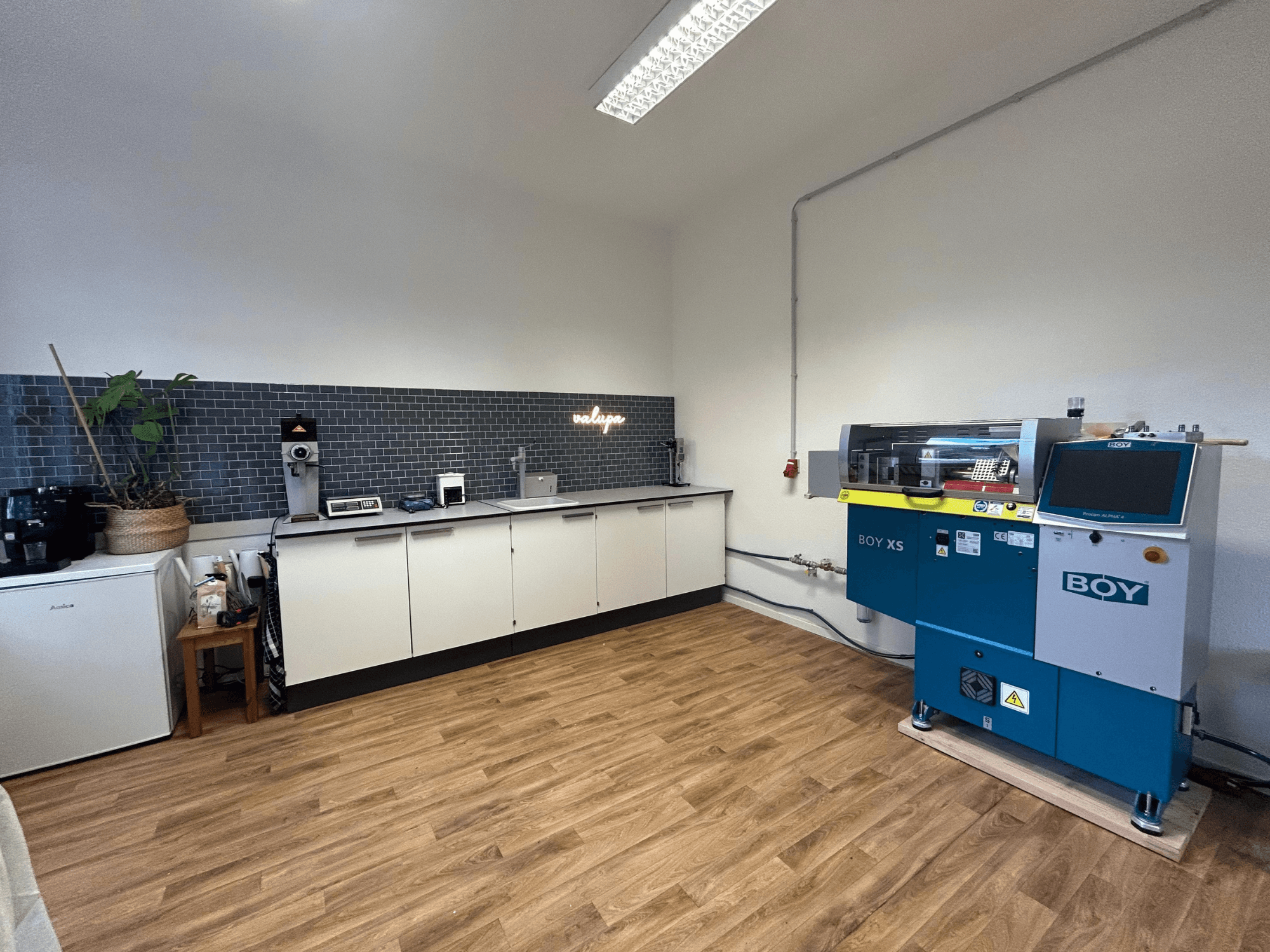 Machine de moulage par injection hydraulique BOY XS 161, vue de face, dans une cuisine moderne avec des appareils électroménagers et un sol en bois.
