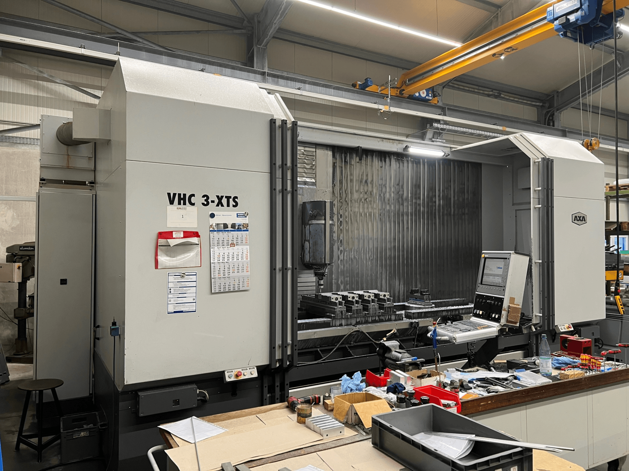 Machine CNC AXA VHC 3-XTS vue de côté, avec panneau de commande et zone de travail encombrée d'outils et de composants.