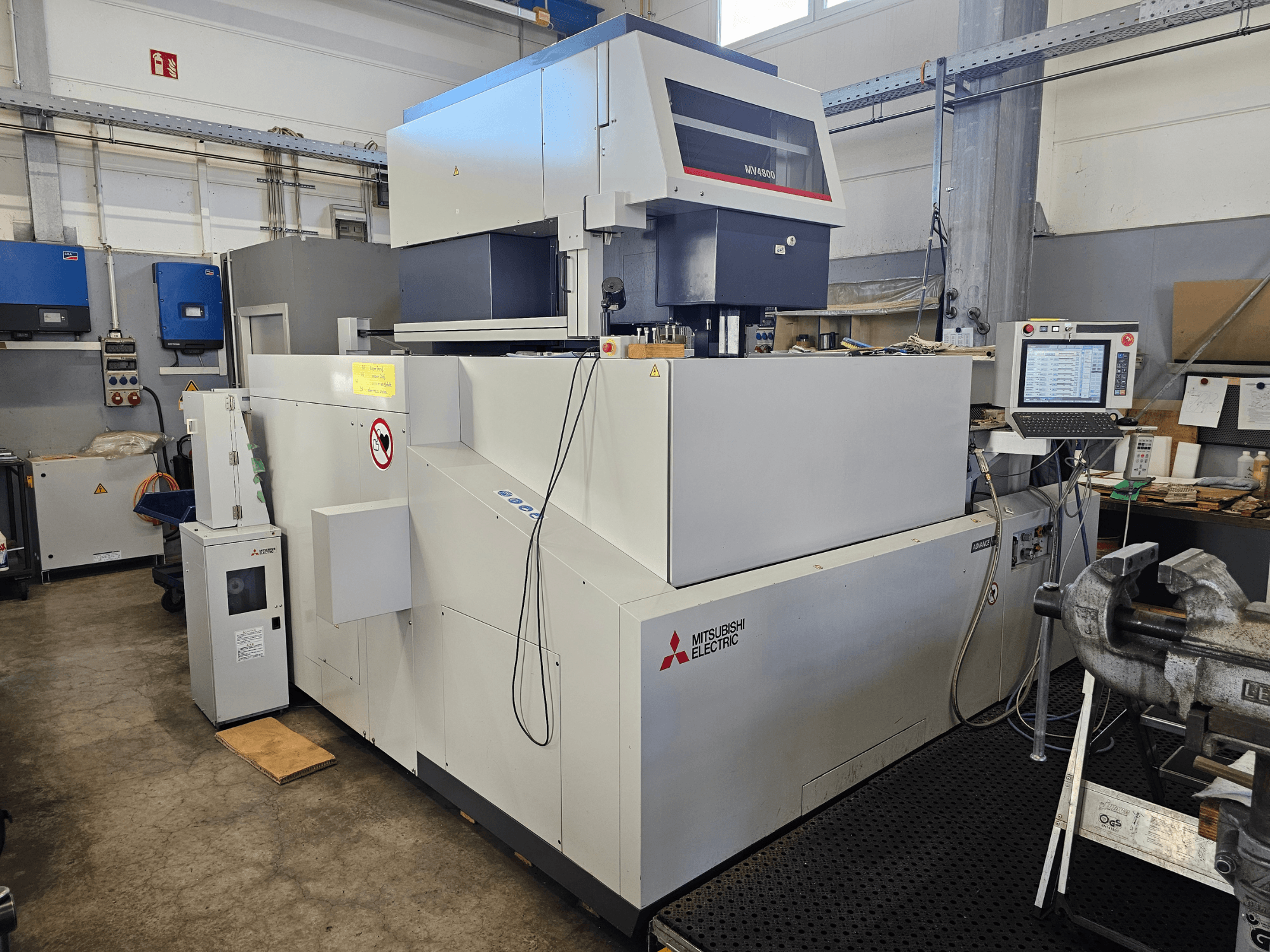 Machine d'électroérosion à fil Mitsubishi MV4800 dans un atelier, vue latérale montrant le panneau de commande, les étiquettes de sécurité et l'équipement environnant.