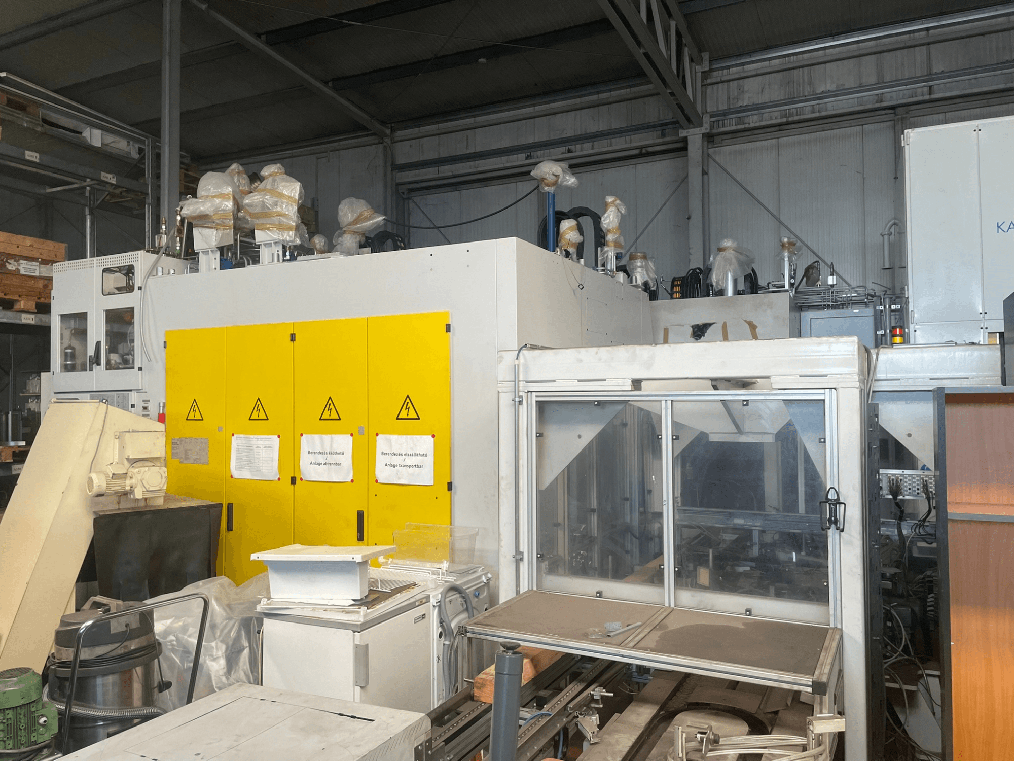 Machine industrielle avec panneau de commande jaune, vue de côté, comportant des étiquettes d'avertissement et des revêtements en plastique, dans un entrepôt.