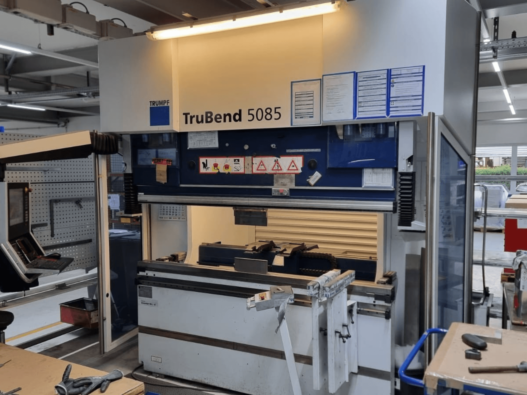 Cintreuse TRUMPF TruBend 5085, vue de face, montrant le panneau de commande, le poste de travail et les avertissements de sécurité à l'intérieur de l'atelier.