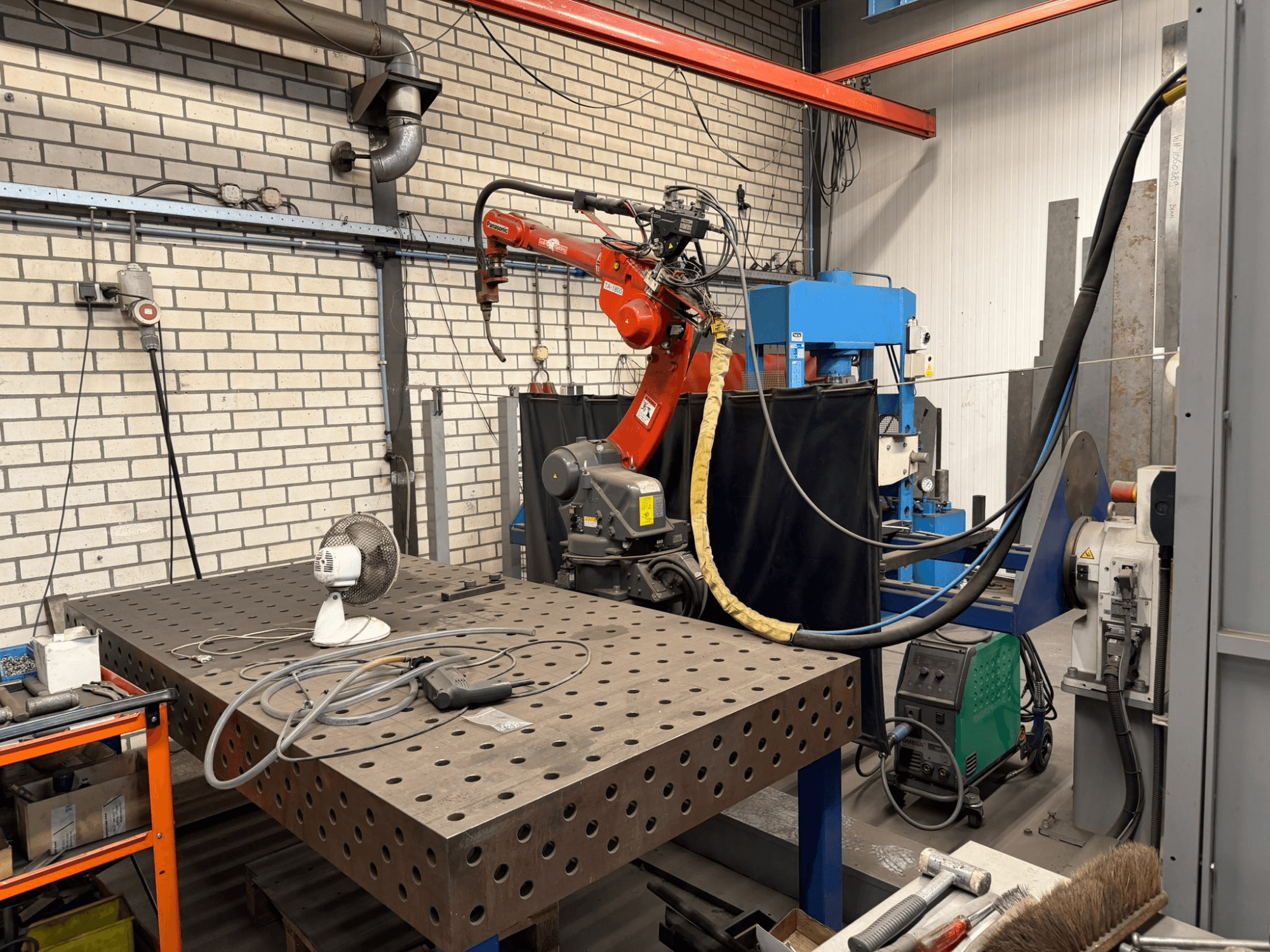 Vue de face d'un robot de soudage PANASONIC TA-1800 sur une station de travail, avec un ventilateur et un équipement de soudage visibles.