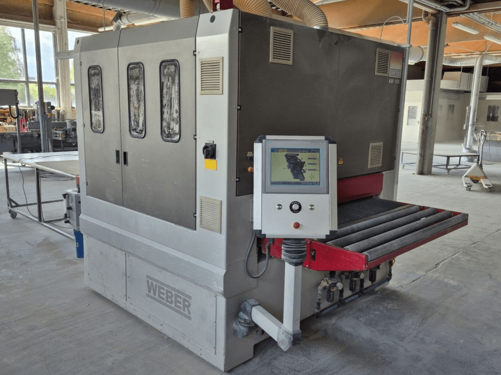 Ponceuse à bande WEBER KSF-3-1350, vue latérale montrant les commandes, le panneau d'alimentation et le convoyeur de ponçage. Environnement industriel avec machines visibles.