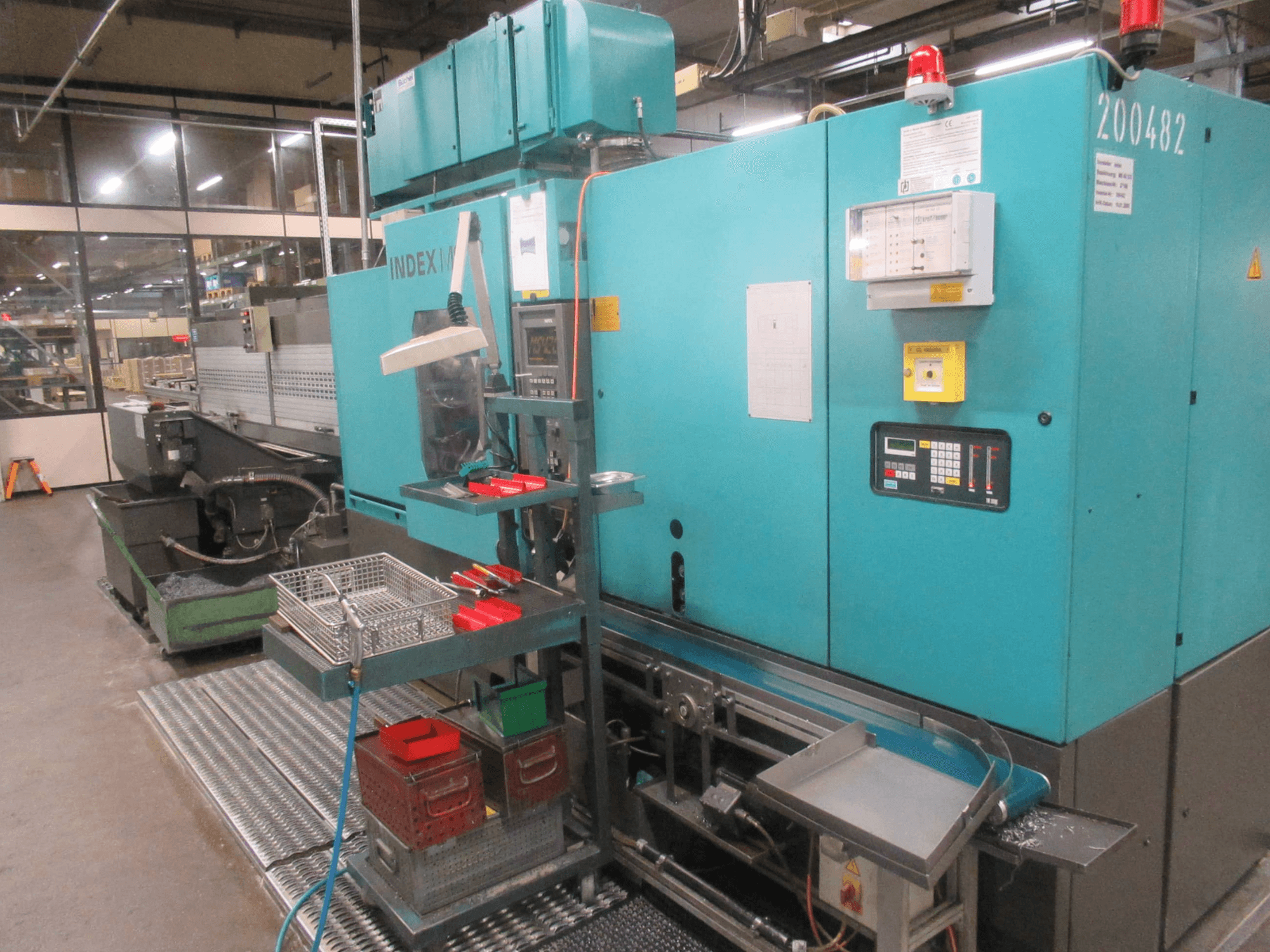 Machine INDEX turquoise dans l'usine, vue de côté ; elle comporte un panneau de commande, un magasin d'outils et un convoyeur. Bacs, tuyaux et alarmes visibles.