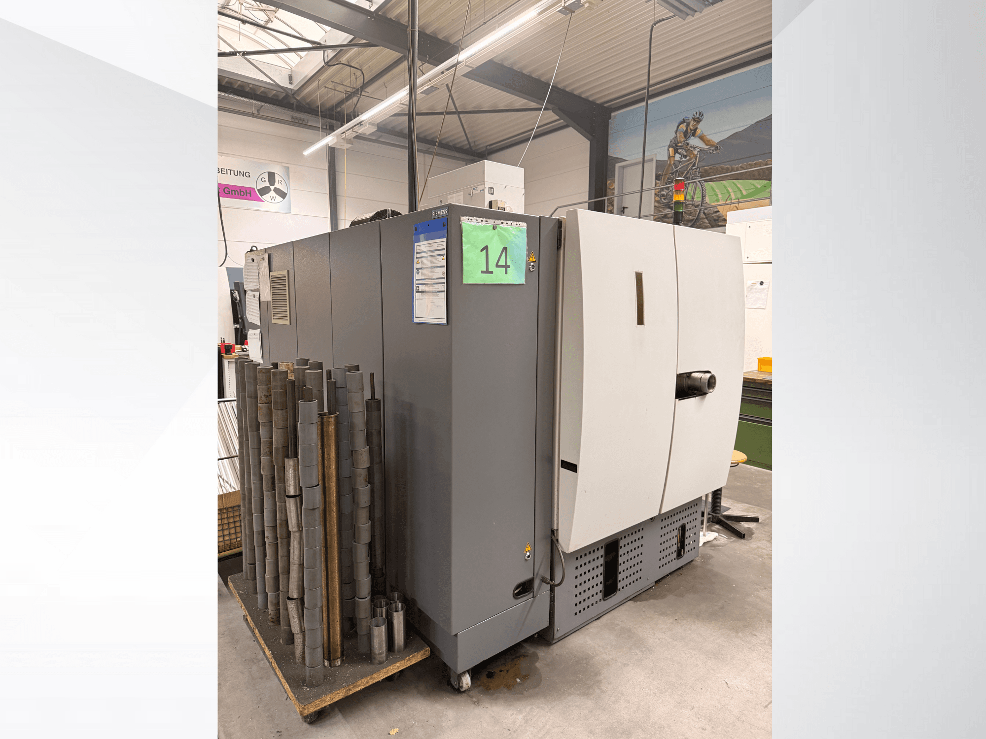 Machine industrielle grise de Siemens, vue de côté, avec un stock de pièces métalliques cylindriques à côté et des étiquettes d'équipement visibles.