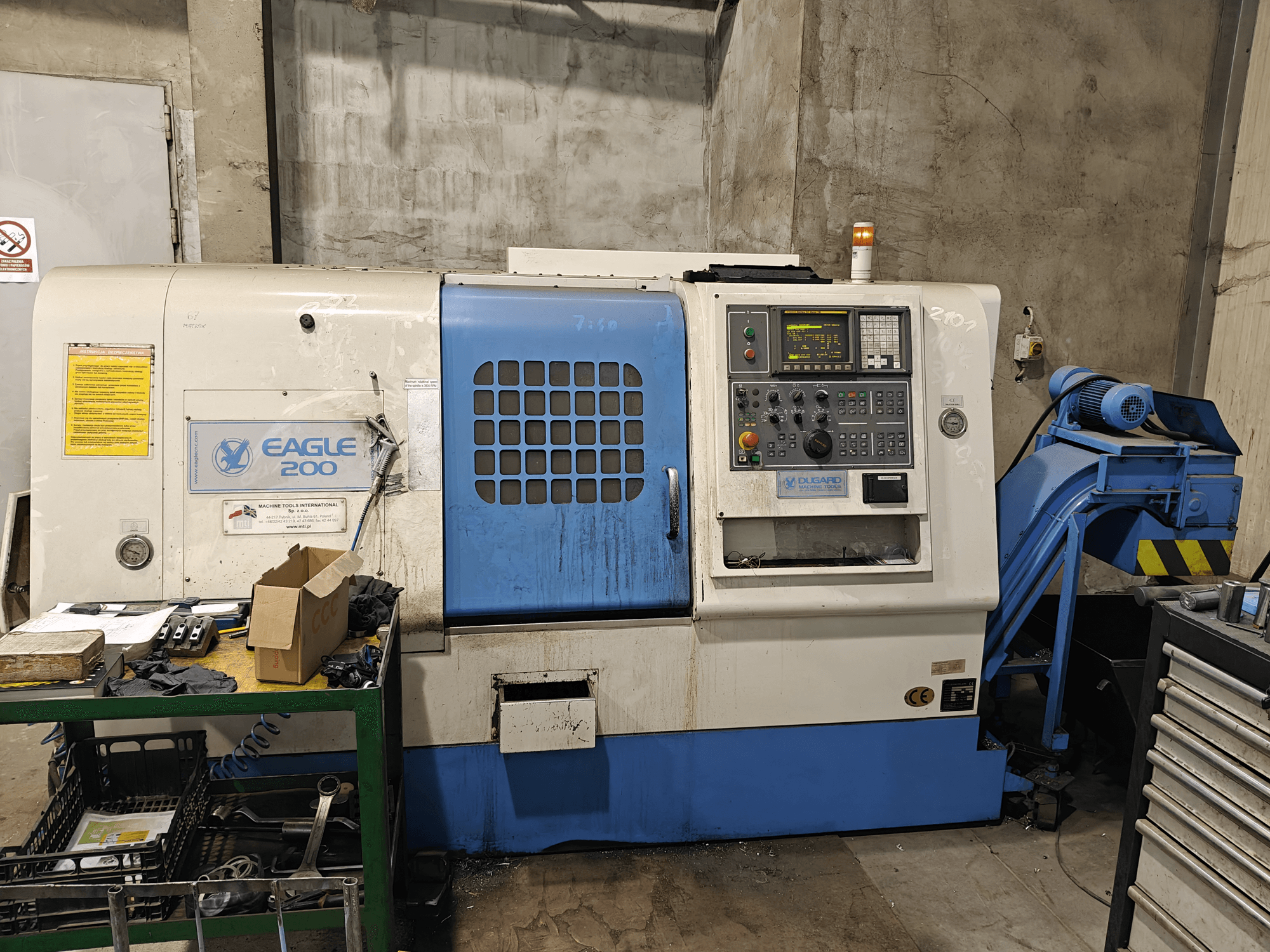 Machine CNC Eagle 200, vue de face avec panneau de commande, divers outils sur un chariot vert. Usure visible et voyants de fonctionnement.