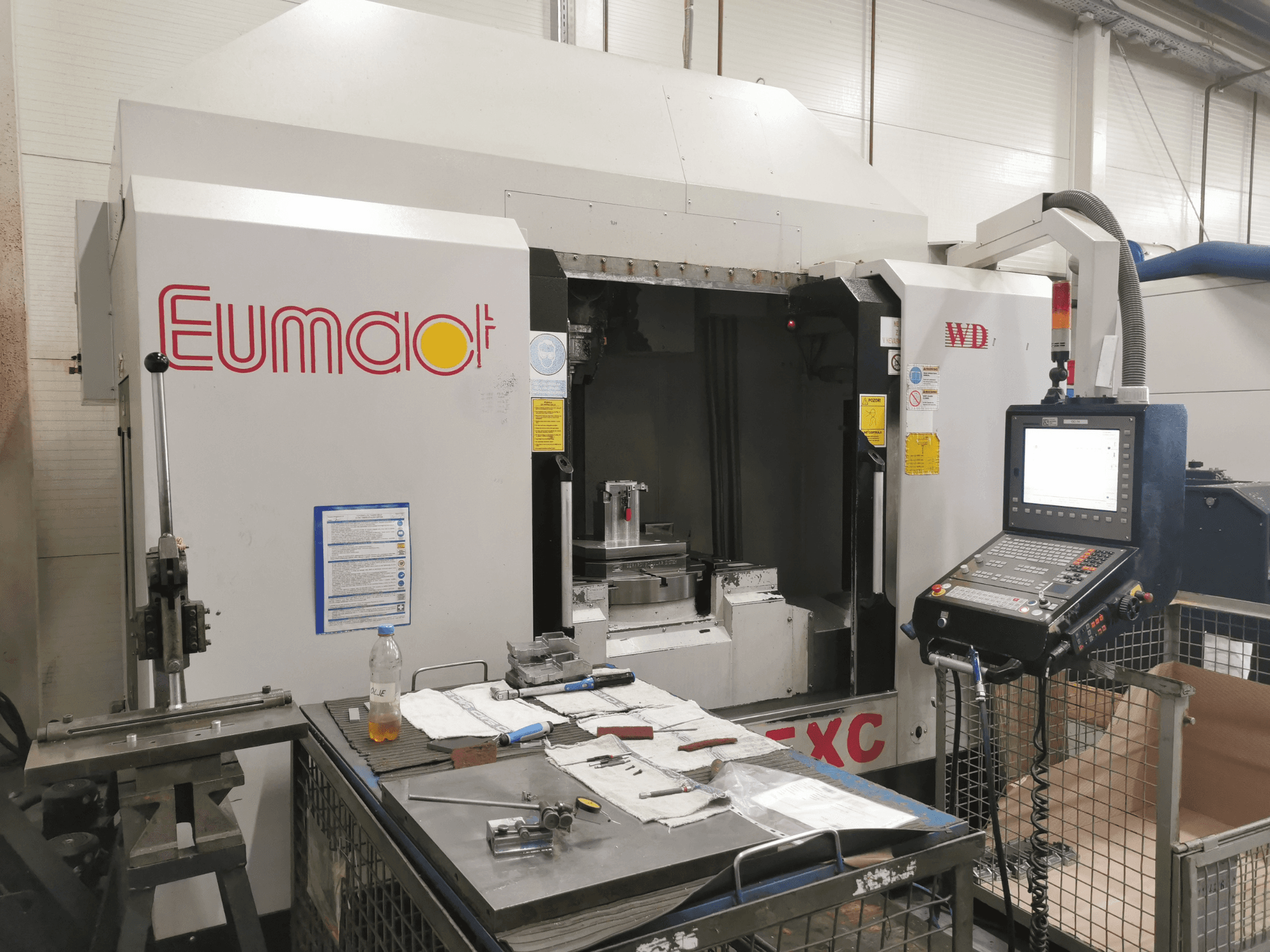 Vue de face d'une machine CNC Eumdaot avec un panneau de commande, des outils et une surface de travail encombrée de composants et de matériaux.