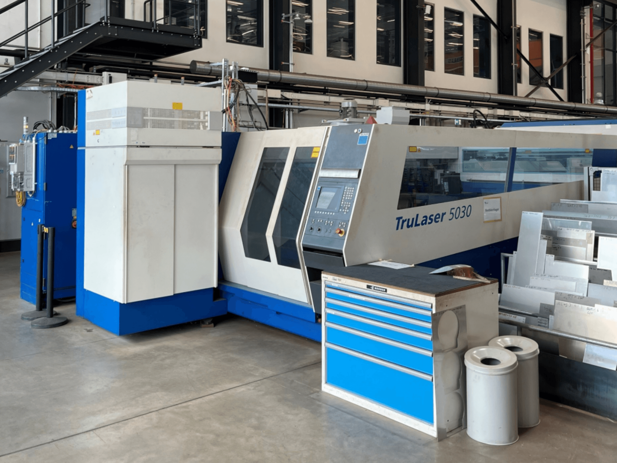 Machine de découpe laser CO₂ TRUMPF TruLaser 5030 vue de face, avec son panneau de commande et l'établi situé à proximité, sur lequel sont posées des tôles.