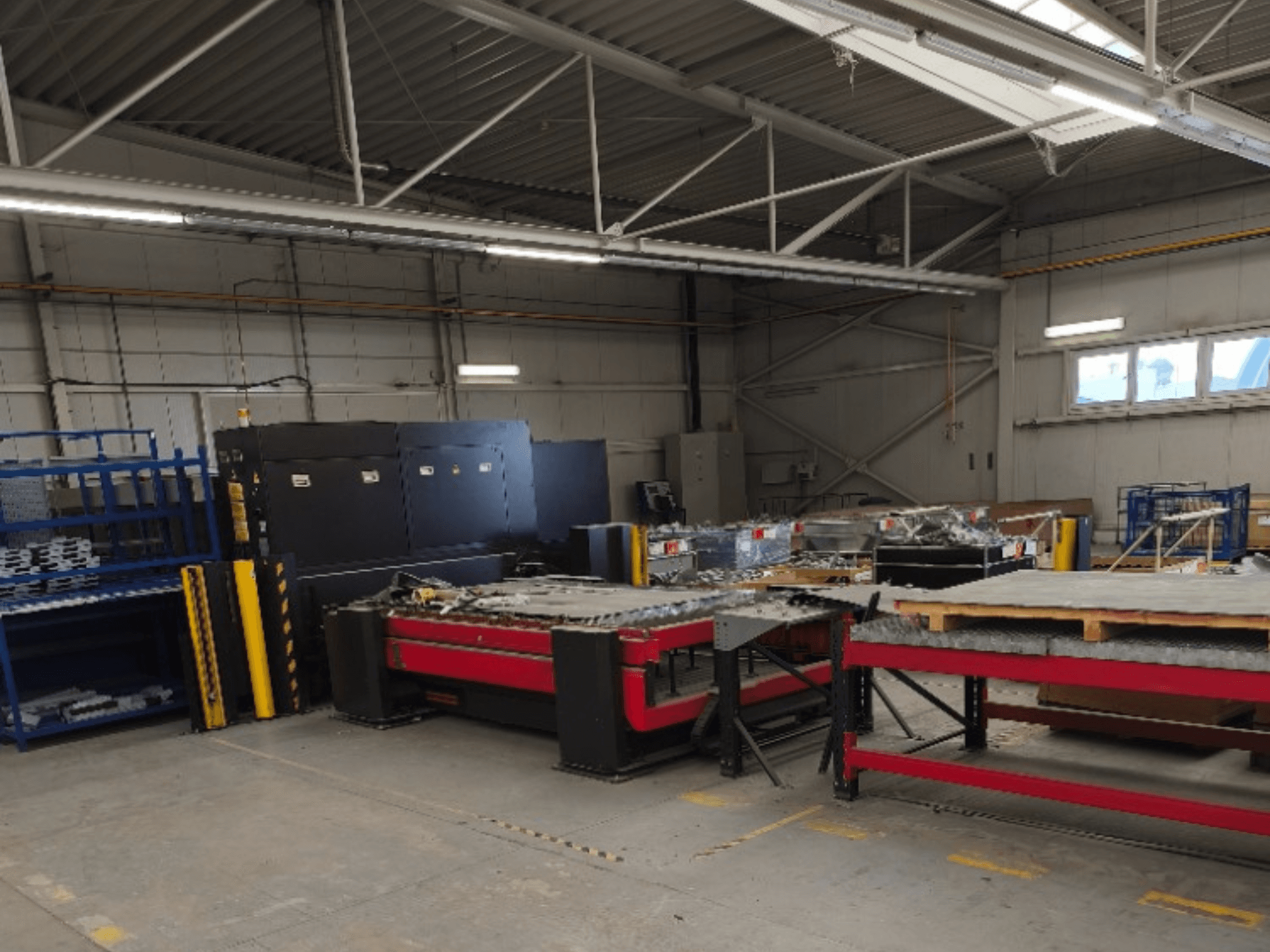 Machine de découpe laser à fibre AMADA LCG 3015 AJ dans un atelier, vue de face, avec des rayonnages de stockage et des matériaux à l'arrière-plan.
