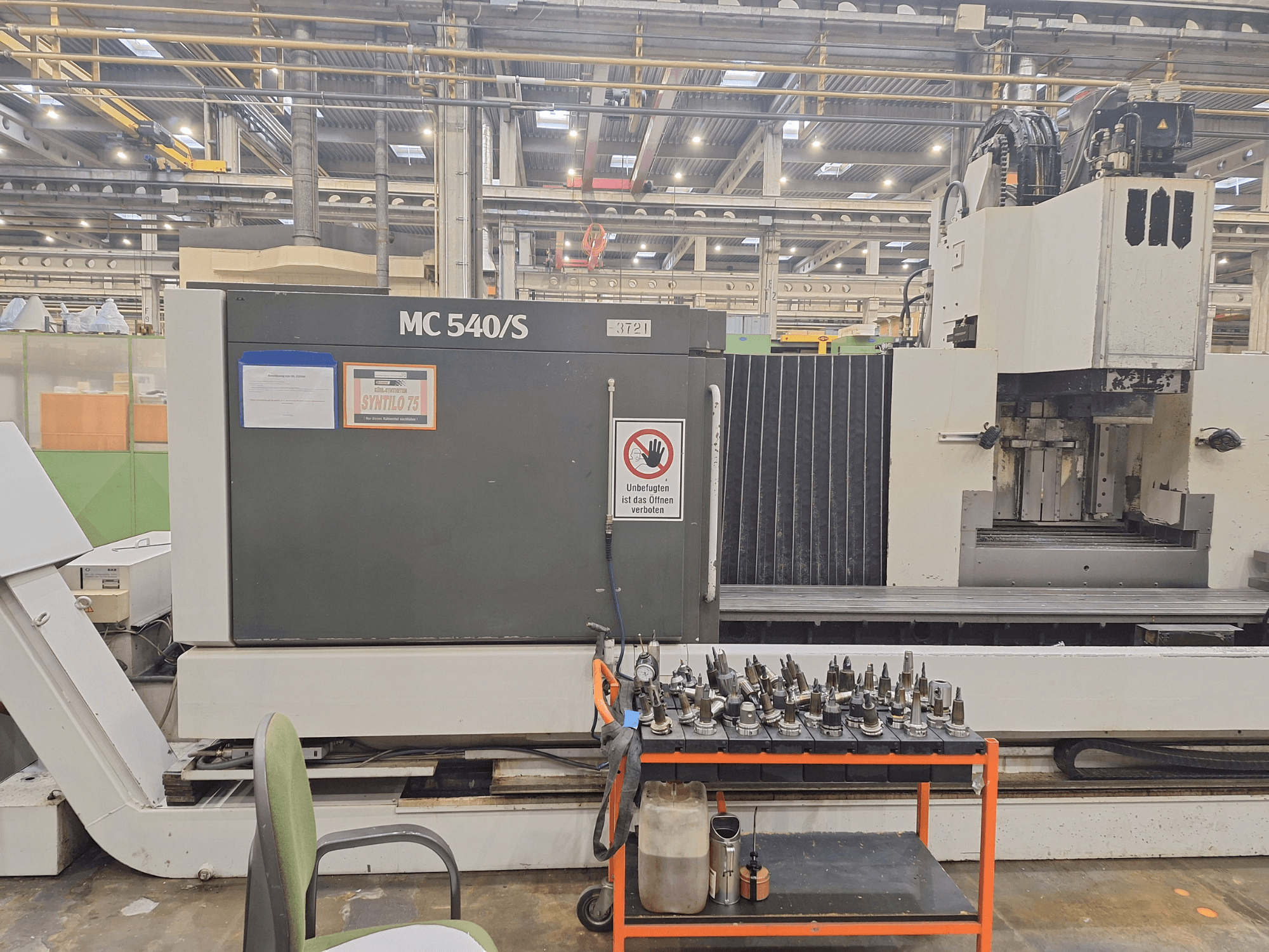 Centre d'usinage vertical STAMA MC 540S dans un atelier, vue latérale avec des porte-outils sur un chariot à proximité.