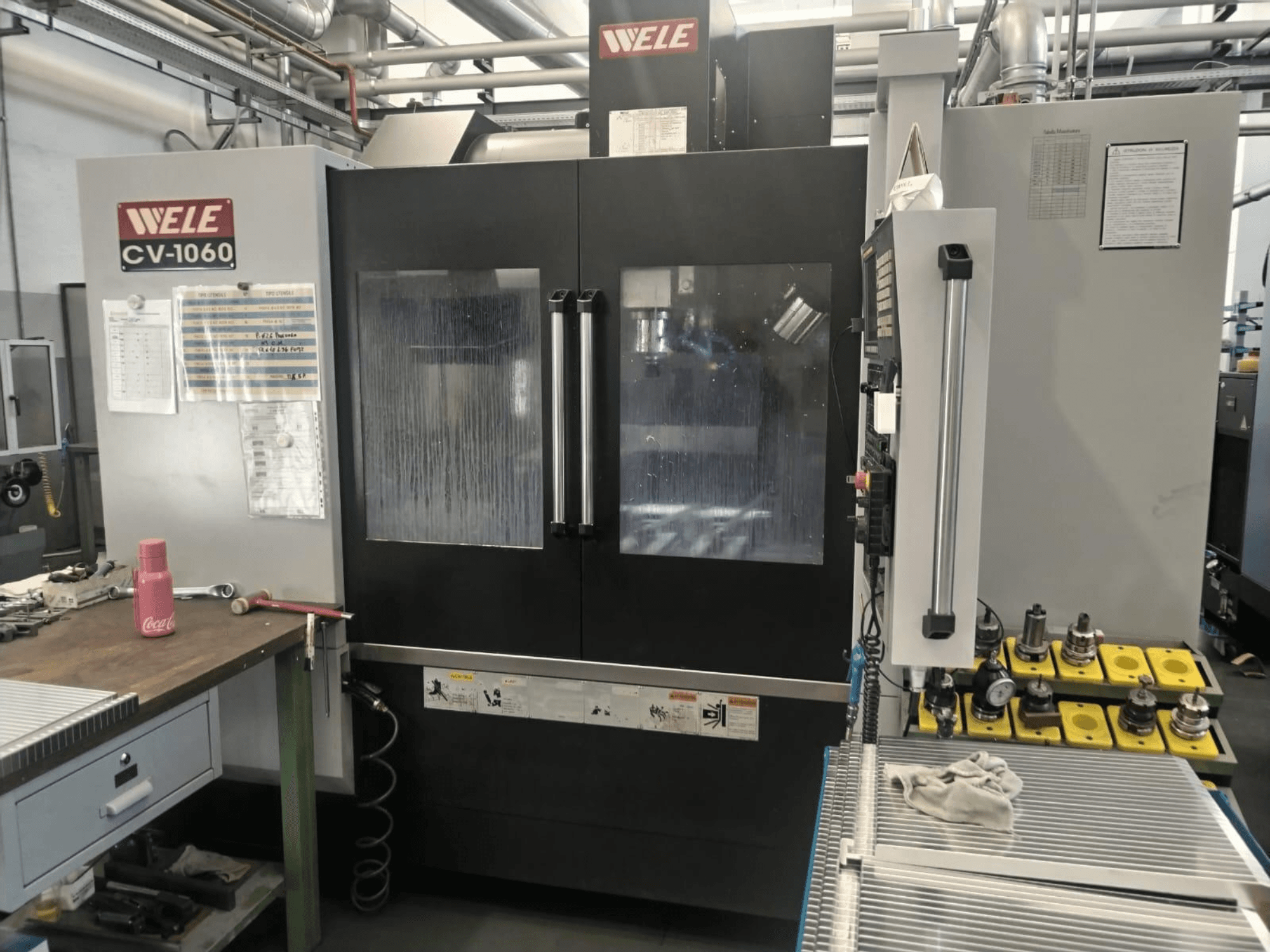 Centre d'usinage vertical Wele CV-1060, vue de face, avec panneau de commande et zone de travail visible ; comprend les outils accessoires et les étiquettes de sécurité.