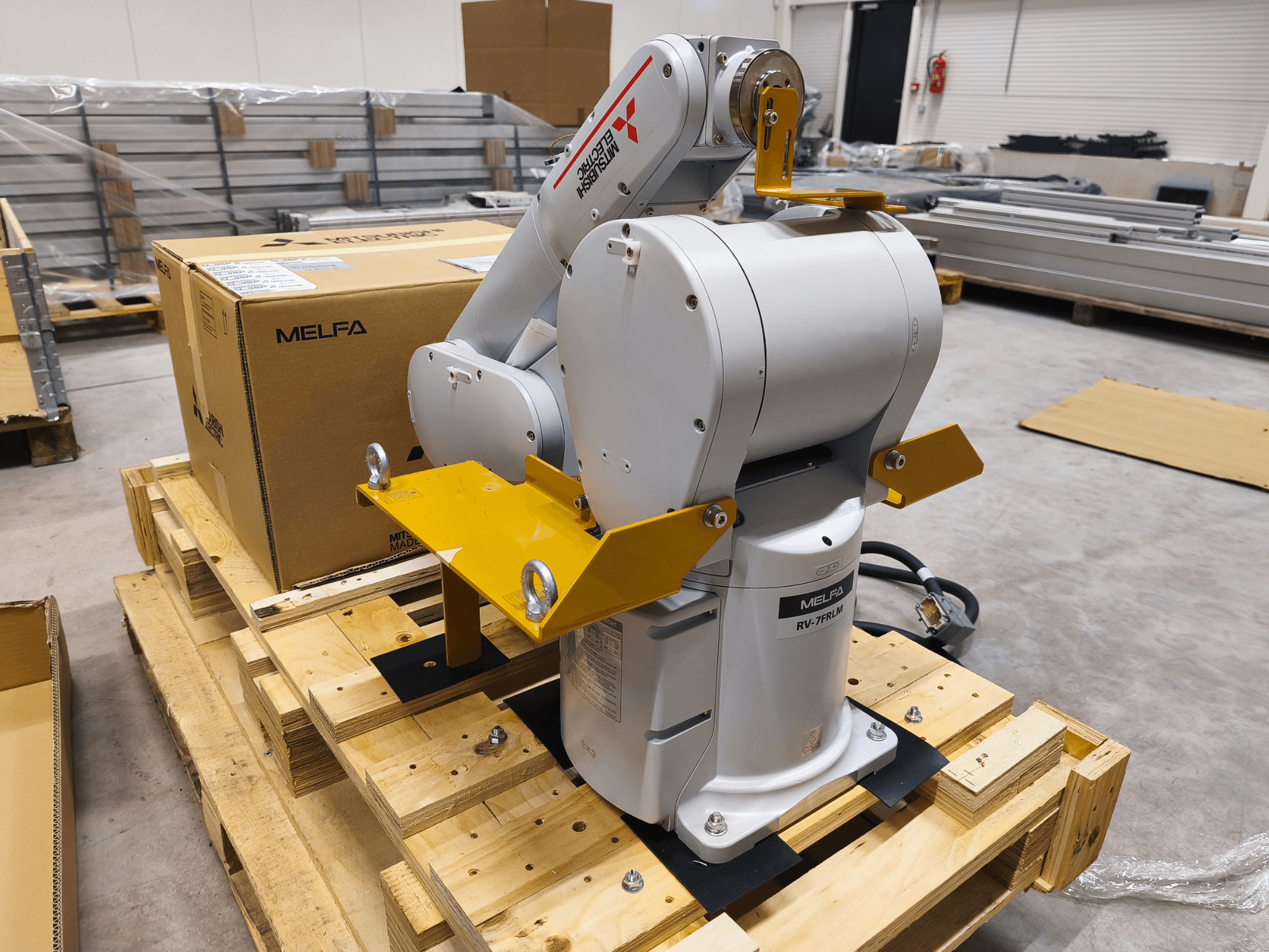 Bras robotique industriel MELFA RV-7FLM sur palette en bois, vue de face, avec supports jaunes et boîte d'emballage grise.