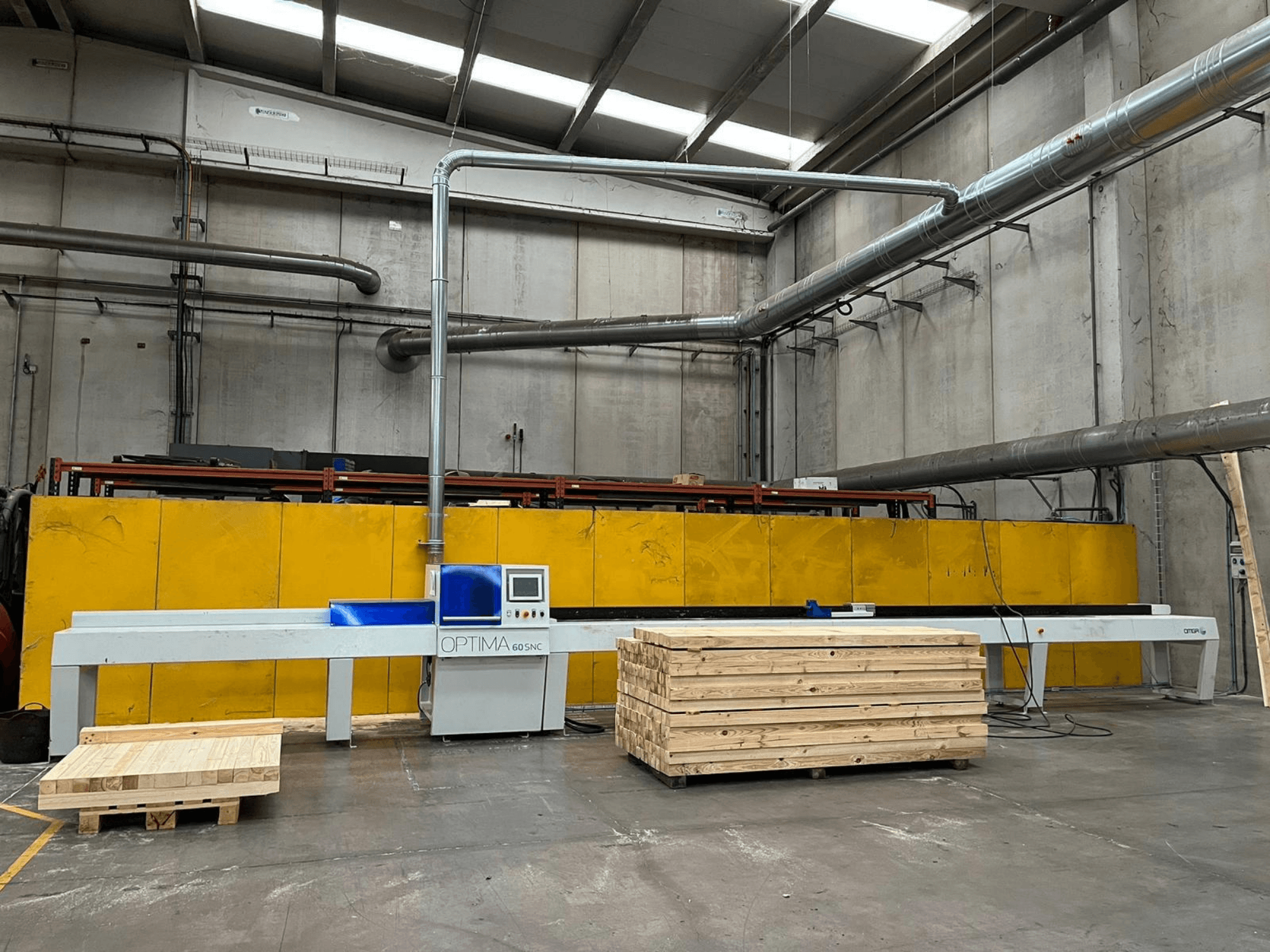 Vue de face de la scie à coupe transversale optimisée OPTIMA 60SNC 6200 CE, avec des palettes en bois et des barrières de sécurité jaunes en arrière-plan.