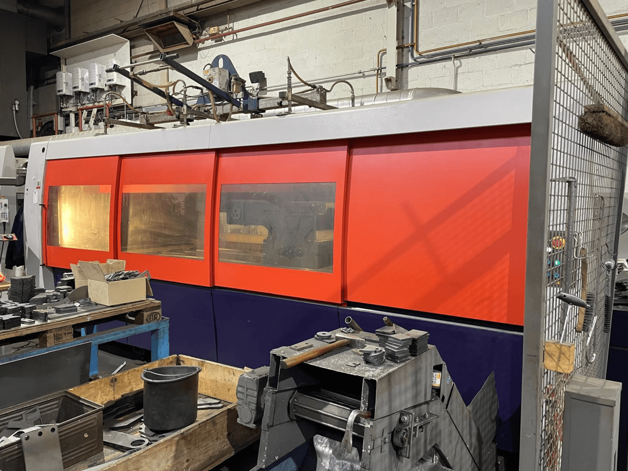 Gros plan d'une machine de découpe laser industrielle orange et blanche, vue de côté, avec panneau de commande et outils d'atelier au premier plan.