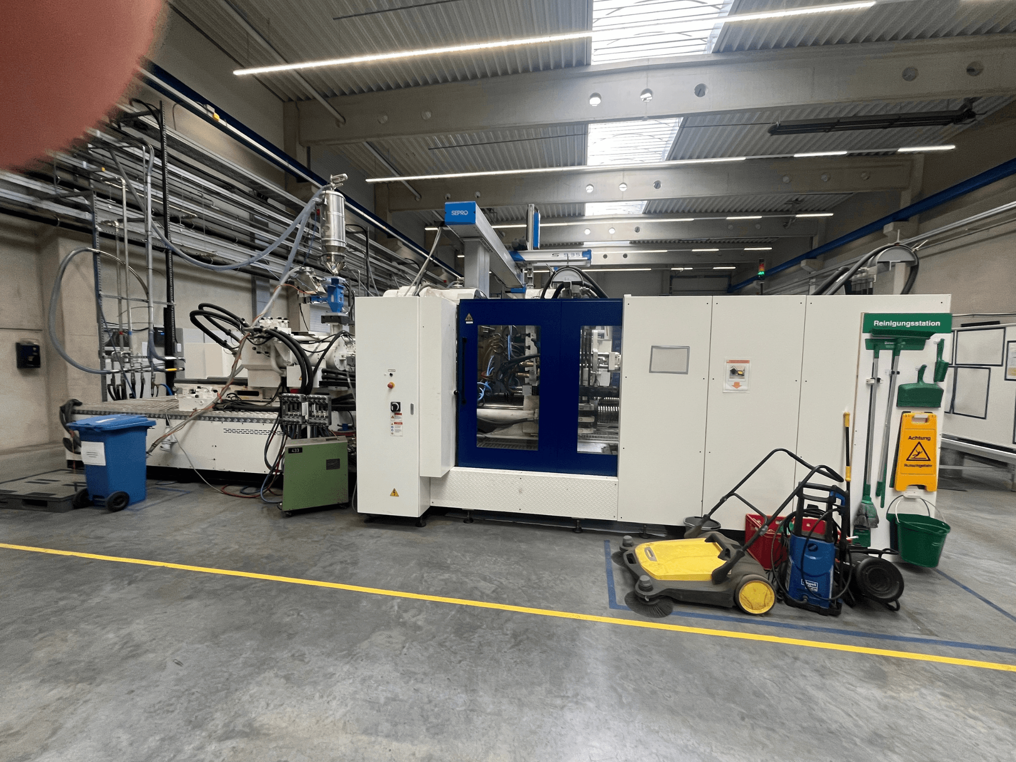 Machine industrielle dans une usine, vue de face, avec des panneaux bleus et blancs, des tuyaux et du matériel de nettoyage à proximité.