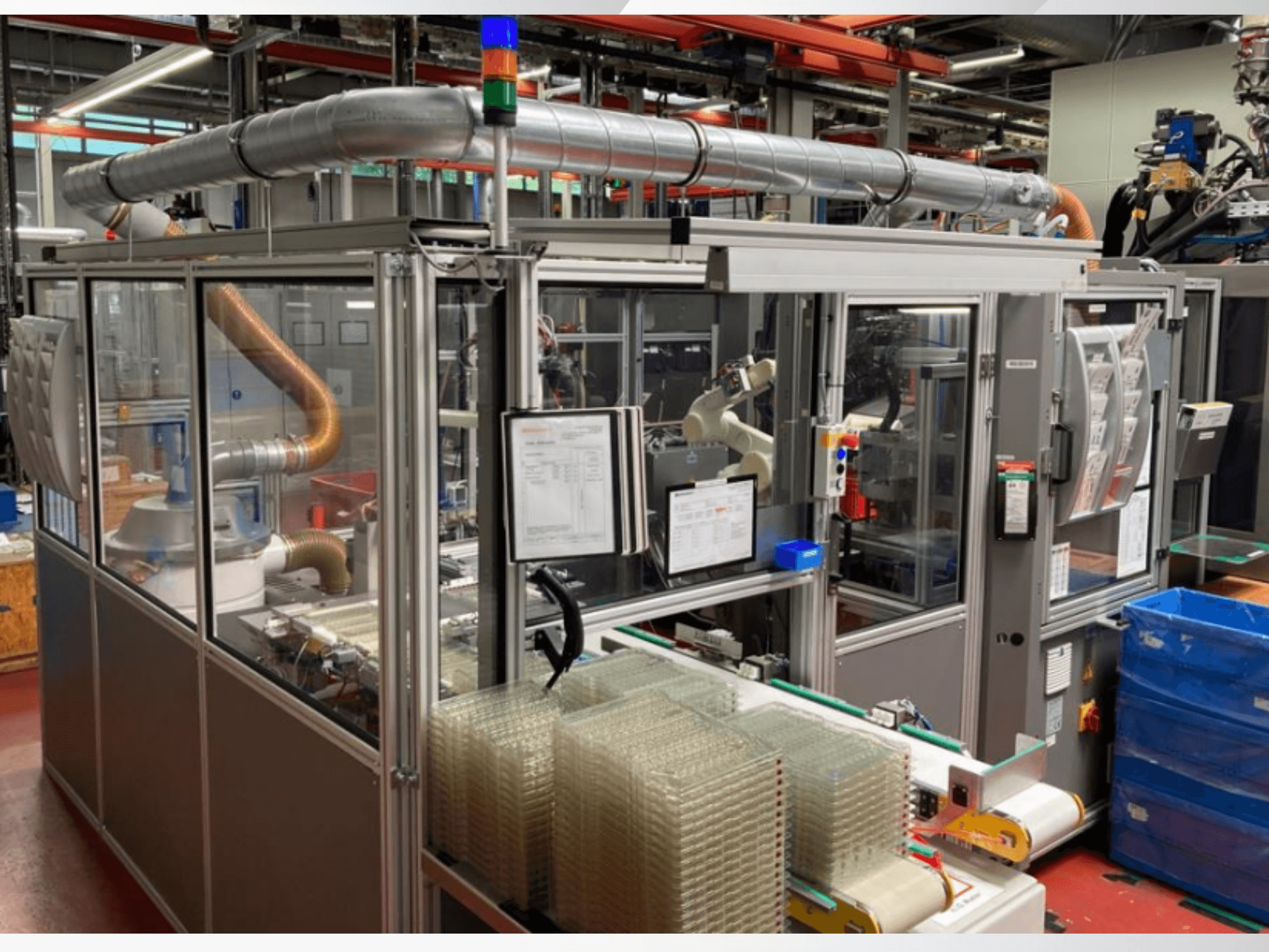 Vue de face d'une machine automatisée avec des tuyaux métalliques, un panneau de commande et des conteneurs en plastique transparent empilés sur le sol d'une usine.