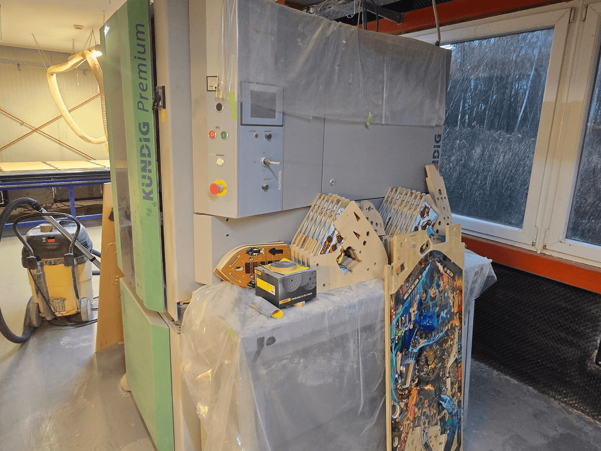 Ponceuse à bande Kundig Premium-1 1100-C dans l'atelier, montrant le panneau de commande et les pièces de bois, vue latérale, avec un aspirateur de poussière à l'arrière-plan.
