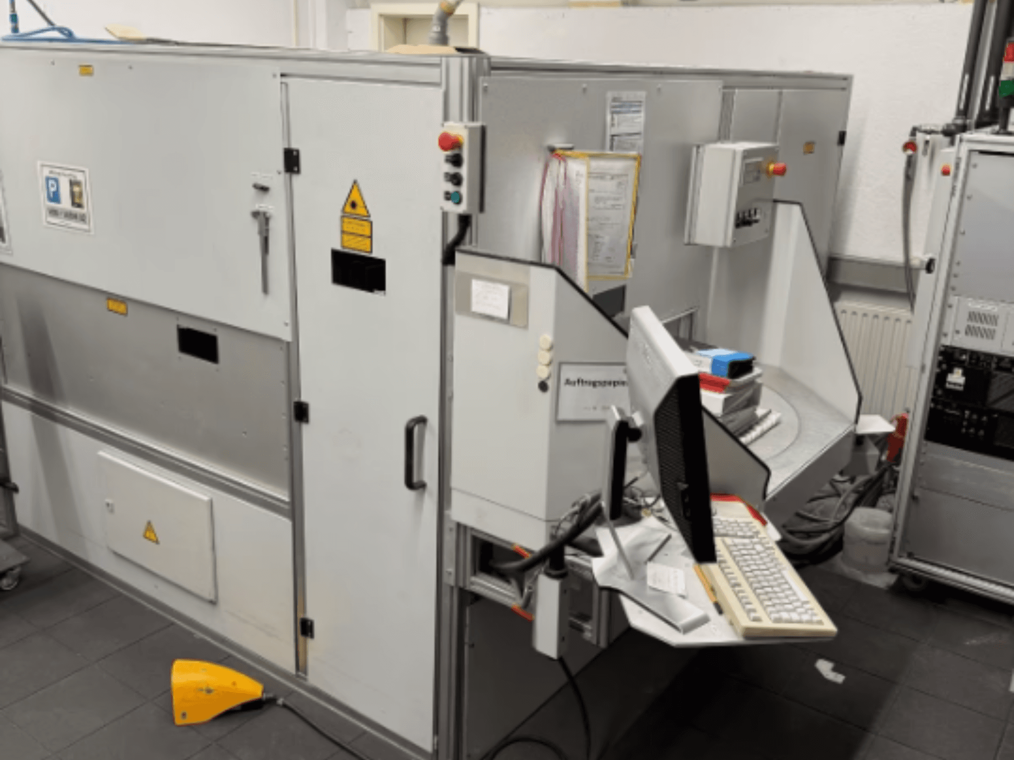 Machine industrielle avec terminal d'ordinateur, vue de côté ; comprend des boutons de commande et des panneaux d'avertissement de sécurité.