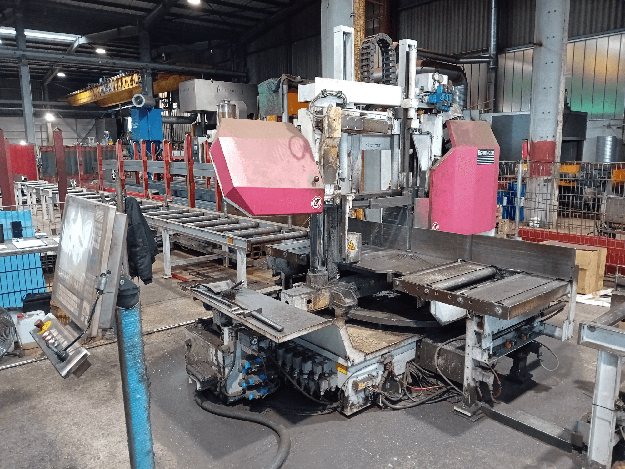 Machine à scier à ruban Behringer dans une usine, vue de face, avec une grande lame de coupe et un panneau de commande avec des boutons.