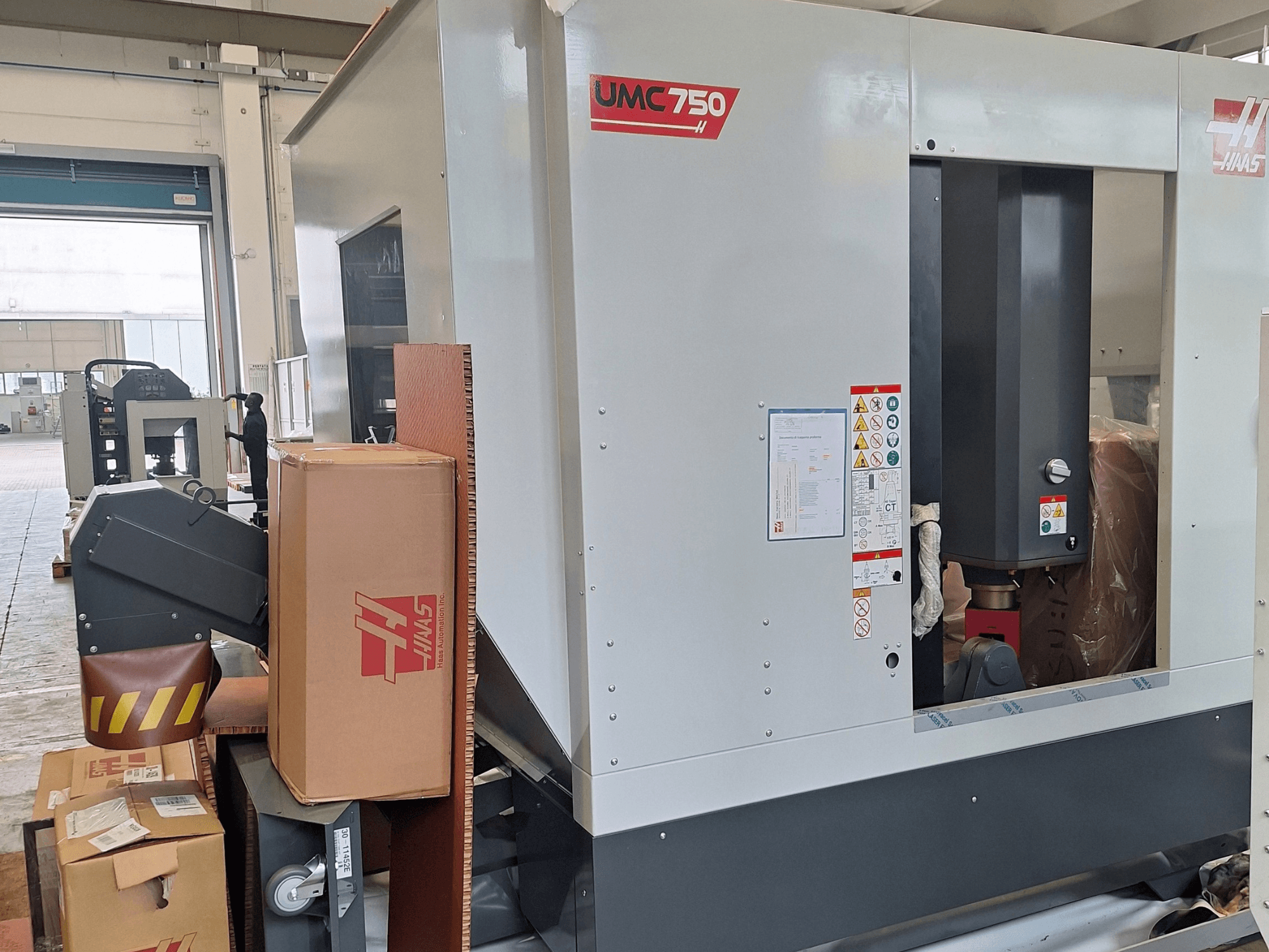 Centre d'usinage universel HAAS UMC-750, vue de profil, présentant le panneau de commande et le rangement des outils, avec étiquettes d'avertissement et emballage.