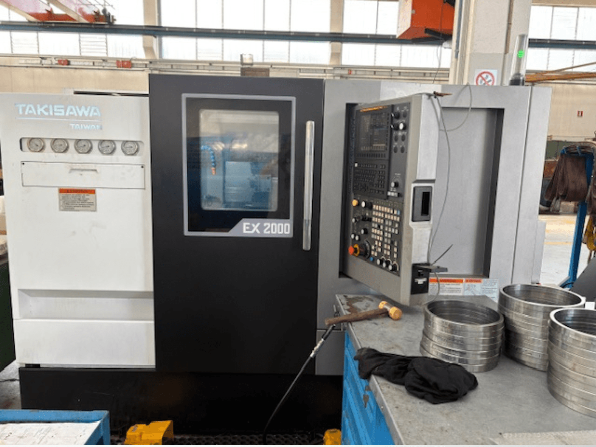 Machine CNC Takisawa EX 2000, vue de face, avec panneau de commande, jauges et pièces métalliques sur une table d'atelier.