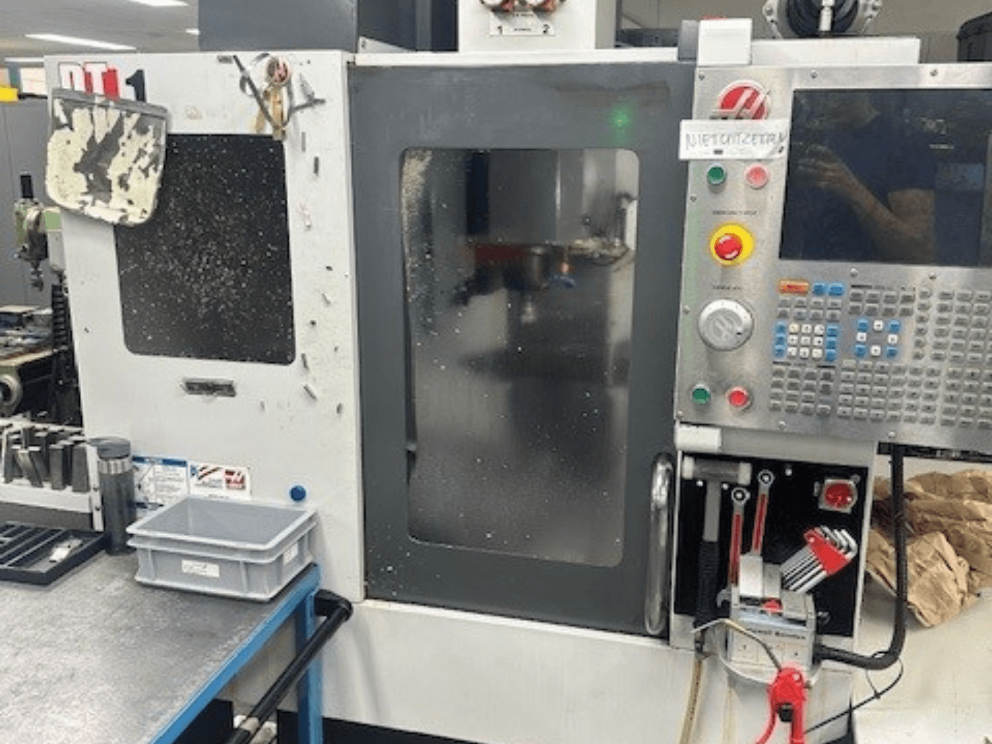Centre d'usinage vertical Haas DT-1, vue de face avec panneau de commande, salissures visibles et plateau d'outils sur l'établi.