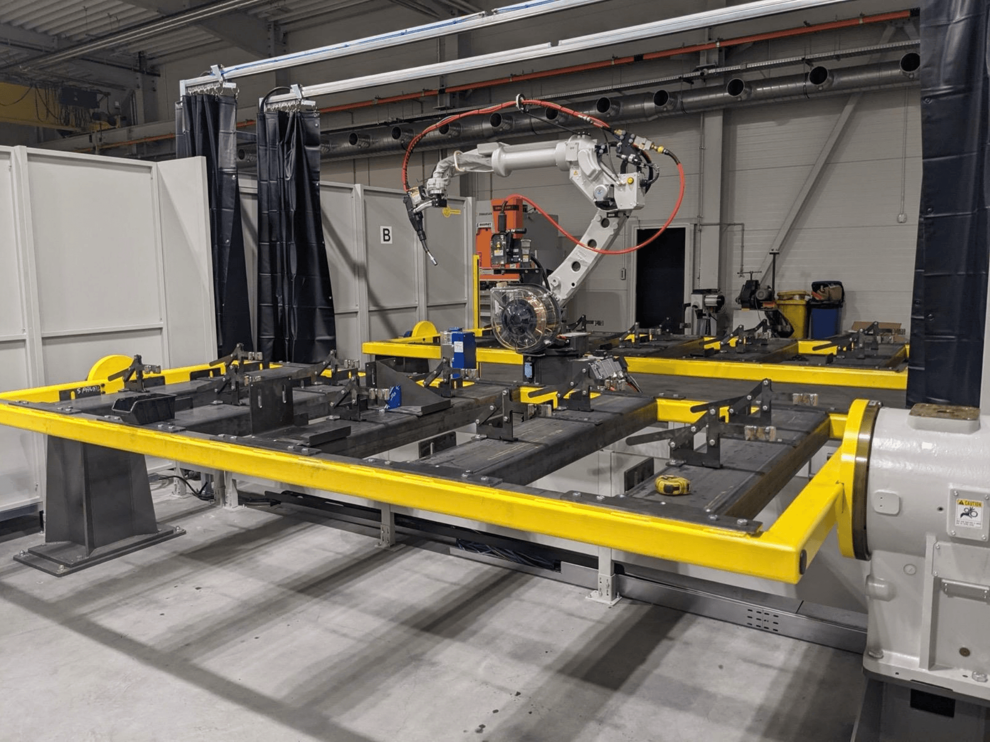 Vue de face du bras robotisé PANASONIC TAWERS TM-2000WG3, placé au-dessus d'une table de travail jaune sur laquelle sont disposées des pièces métalliques destinées à être assemblées.